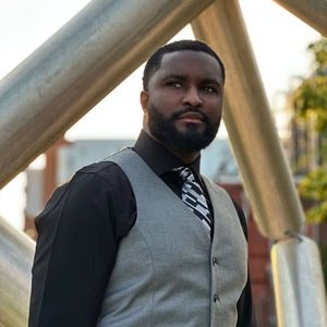 A man with a beard wearing a gray vest, black shirt, and striped tie, standing outdoors near metallic structures with trees in the background.