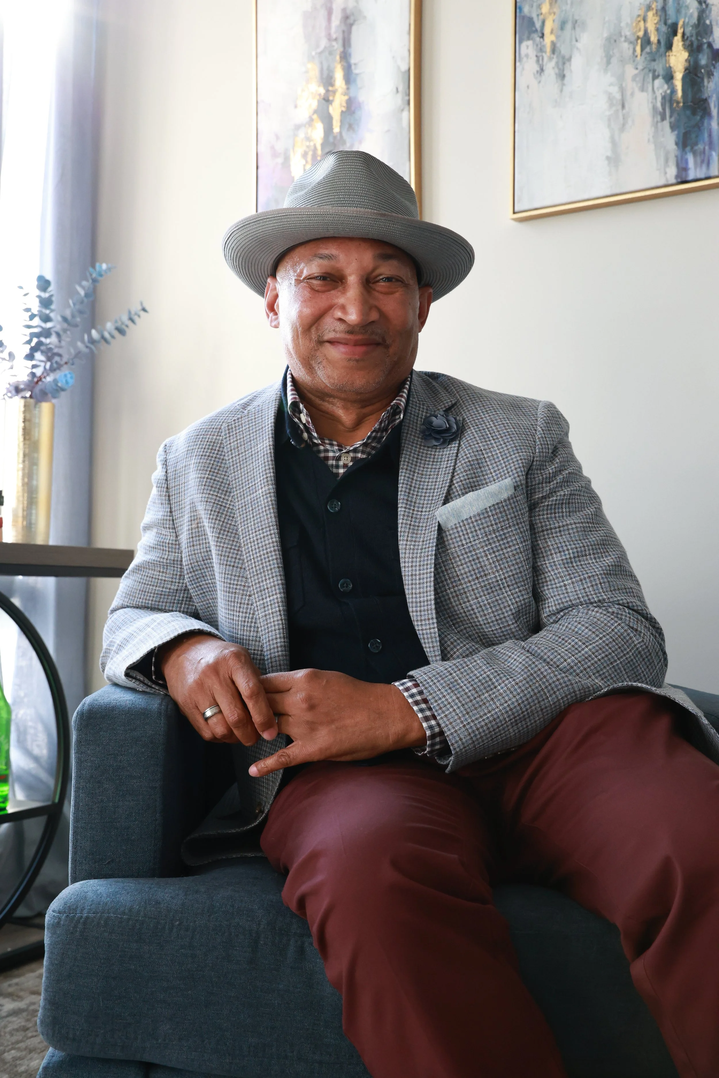 A man sitting on a gray couch, wearing a gray hat, a checkered blazer, a dark shirt, and maroon pants, smiling at the camera, with modern art on the wall behind him.
