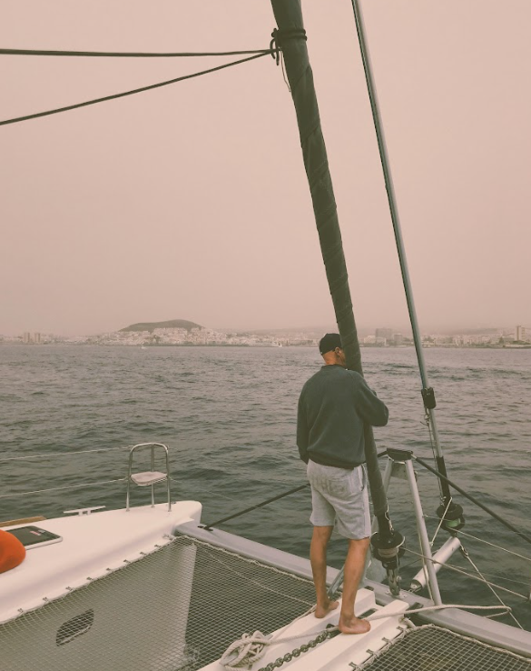 Man standing on a boat deck, looking at the water, with a city and island in the background, under overcast sky.