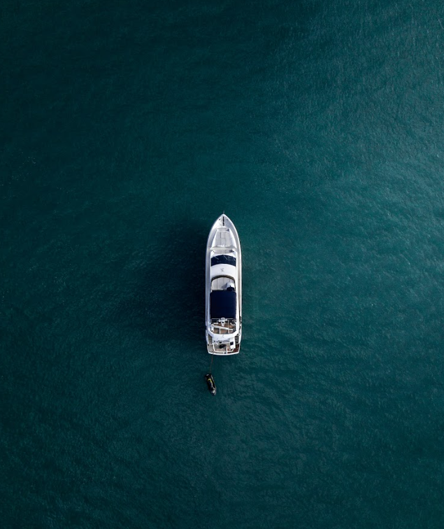 An aerial view of a yacht floating on calm blue water, with a small motorboat attached at the stern.