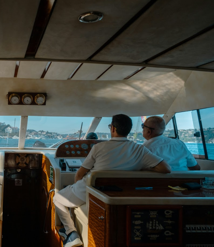 Two men sit in the bridge of a boat, steering it through a body of water with a cityscape in the background during daytime.