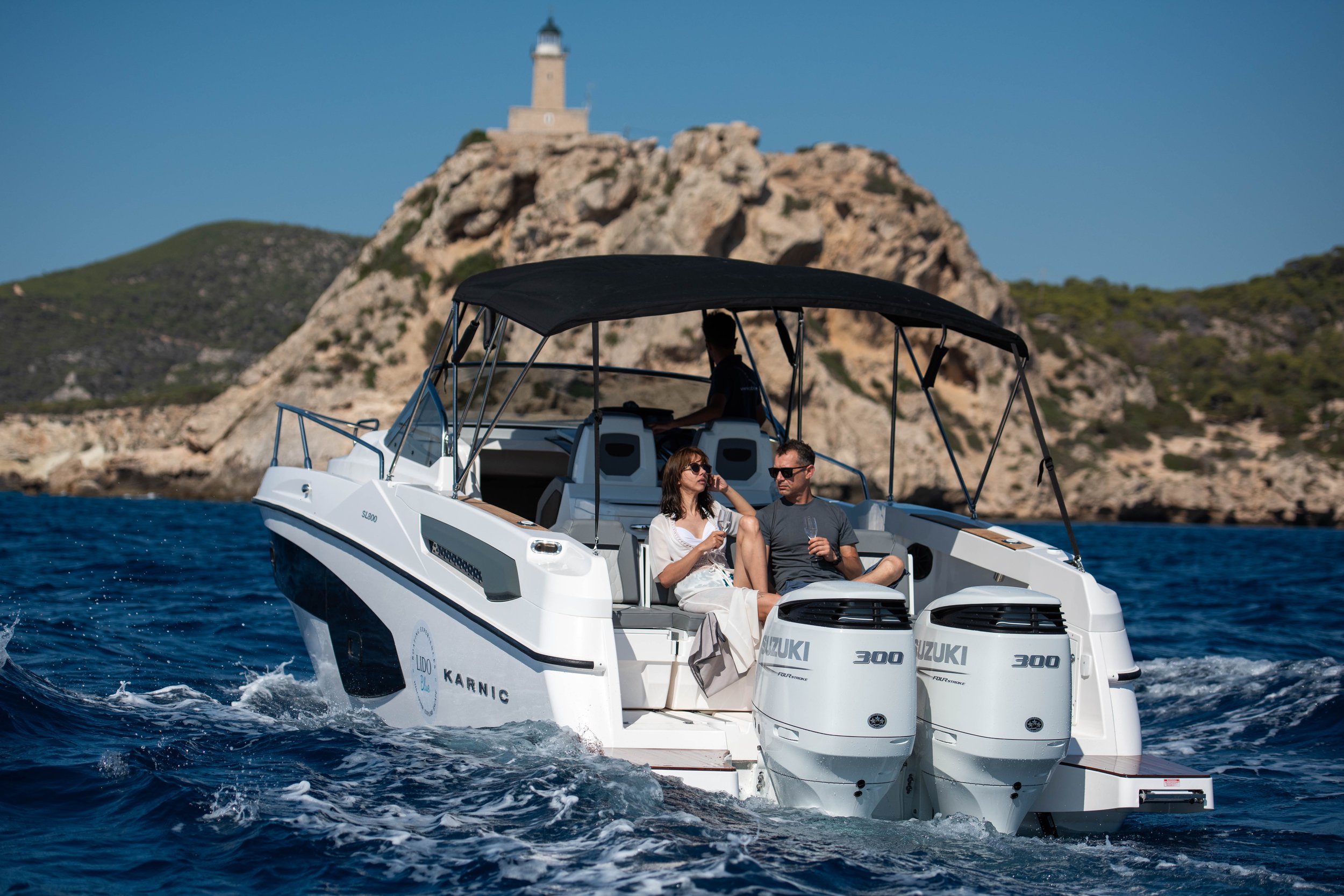 Two people sitting on a white motorboat with twin outboard engines, sailing on the water with a rocky island and lighthouse in the background.