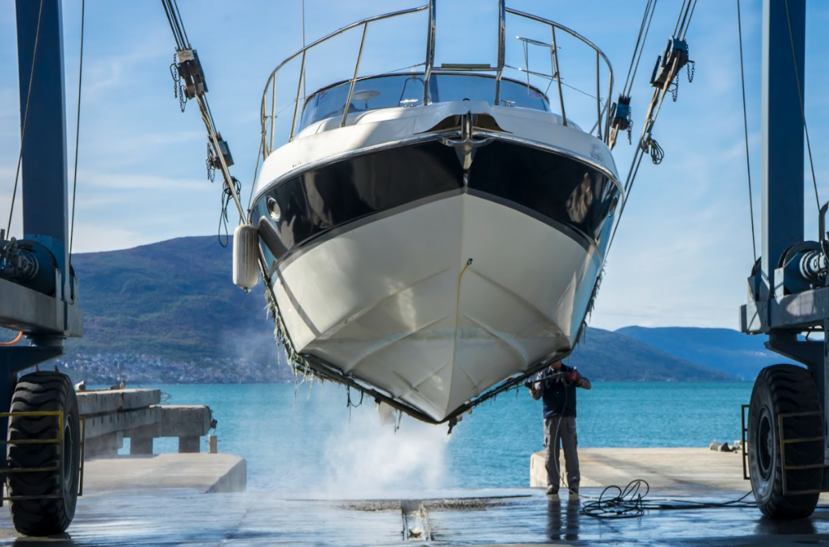 A boat being lifted out of the water by a boat lift at a marina with mountains and water in the background.