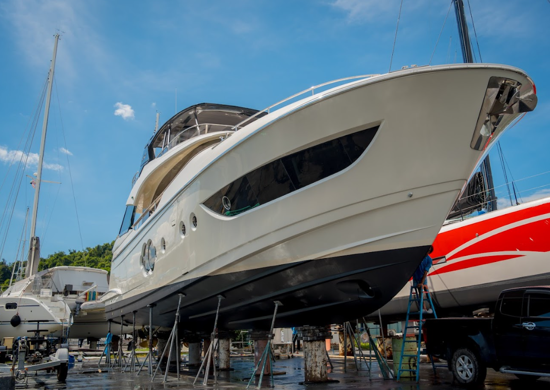 A large white yacht on land, supported by metal stands, with a black hull bottom and a ladder leaning against it. There are other boats and a red and white boat nearby.