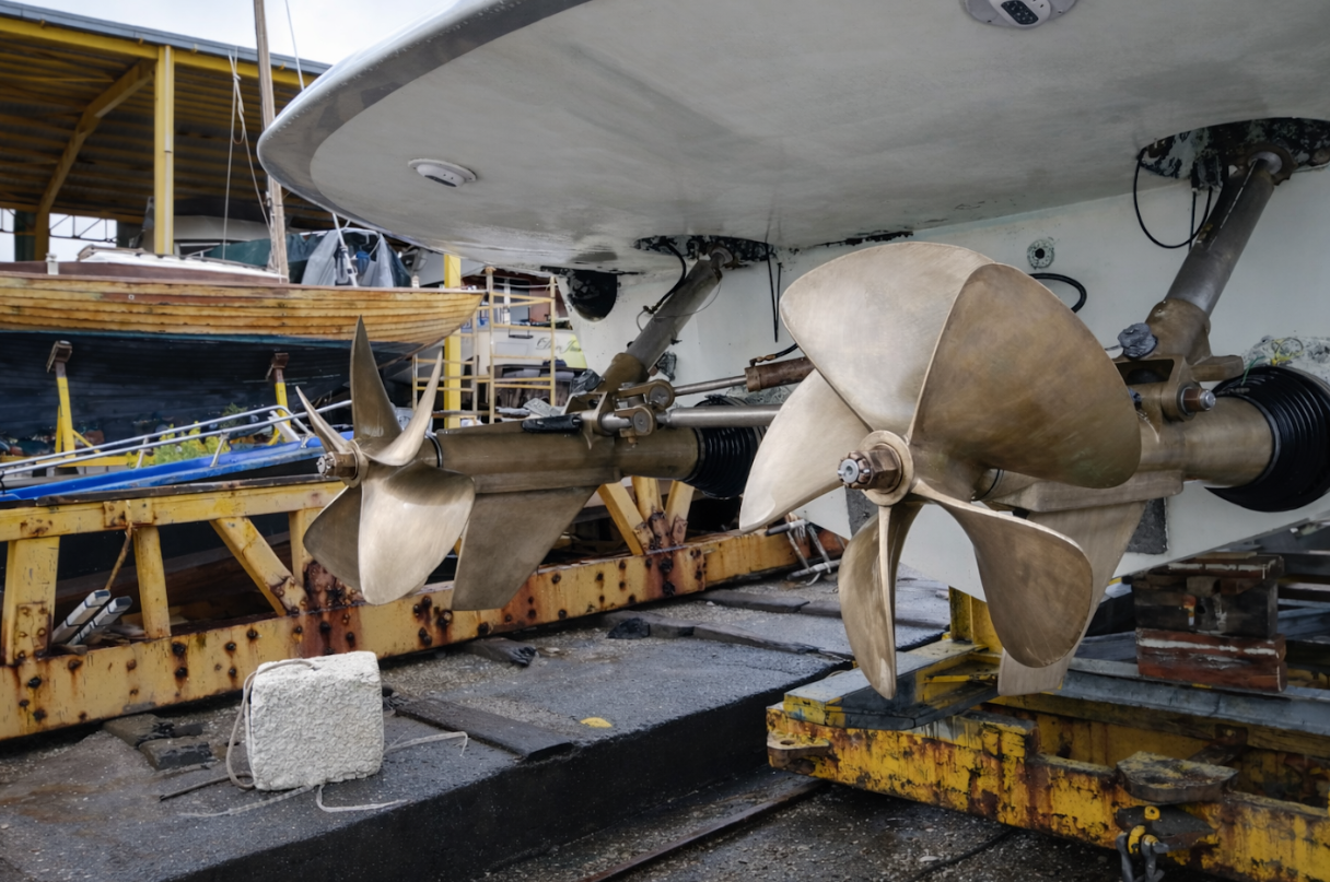 Close-up of a boat's outboard motor with two large brass propellers, situated on a dock with other boats and boat parts in the background.