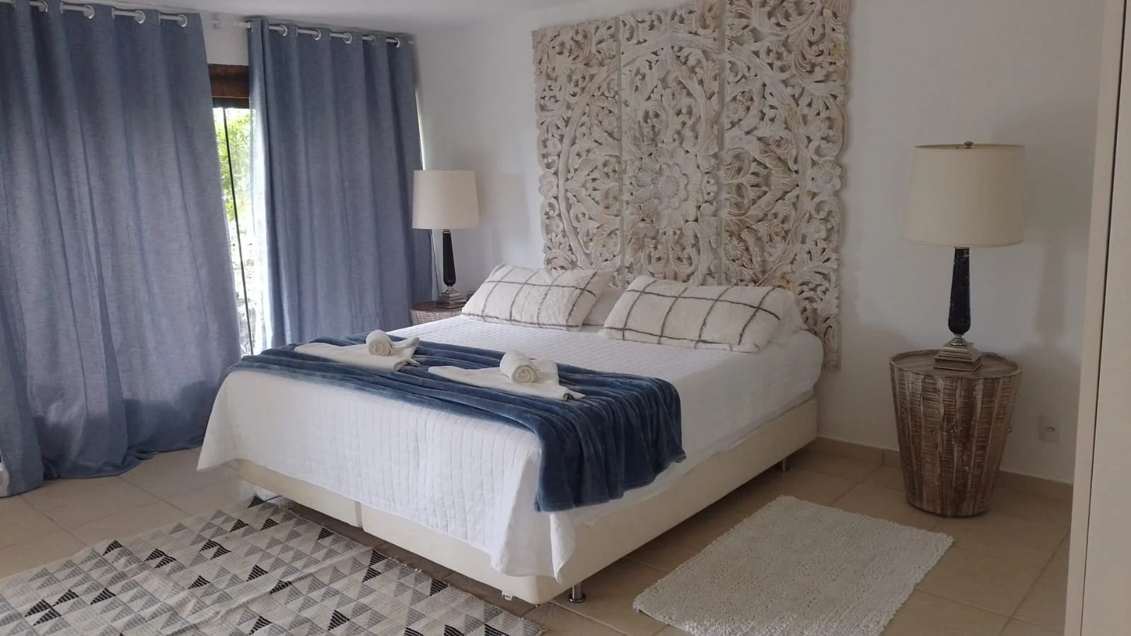 A neatly made bed with white linens and two matching pillows, topped with a dark blue throw blanket and two rolled towels, in a bedroom with a large carved wooden headboard, two side tables with lamps, blue curtains, a tile floor, and a small cream r