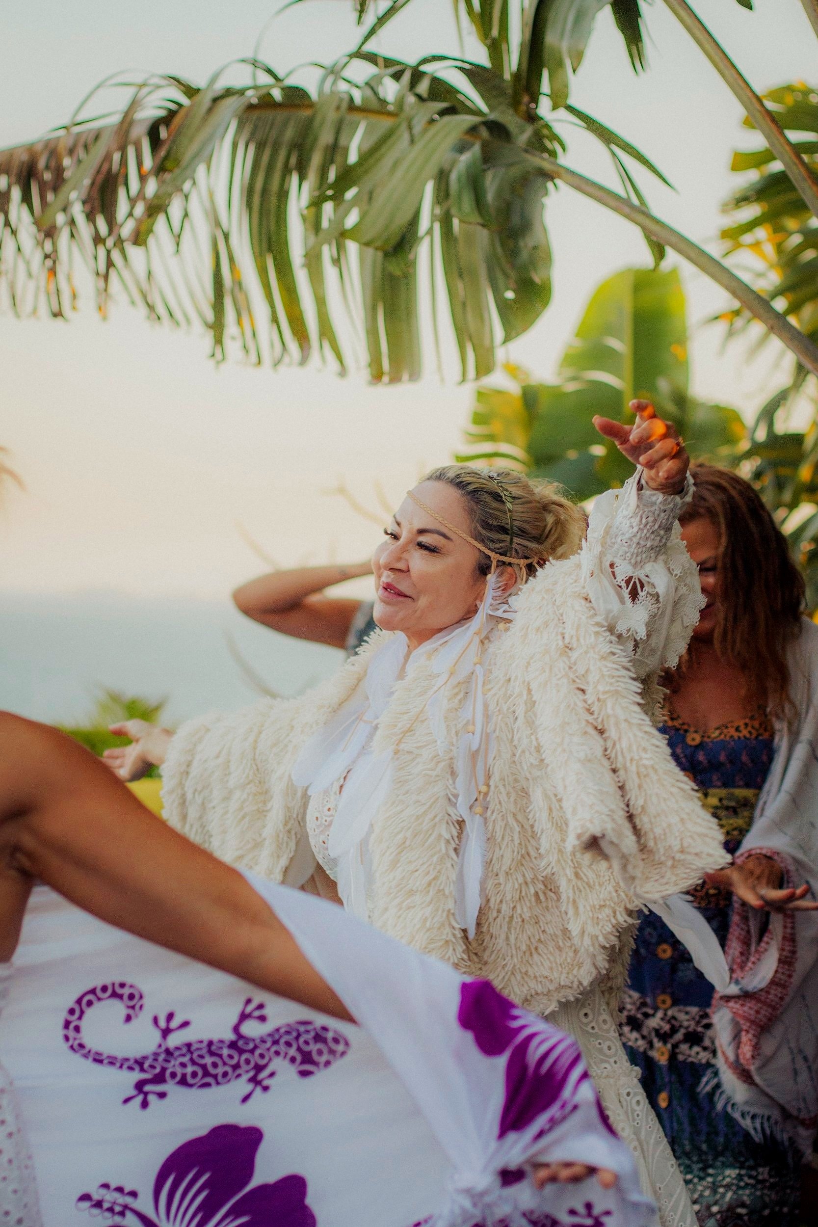 A woman with blonde hair dancing outdoors among tropical plants, wearing a cream-colored fur coat and a headband, smiling with eyes closed, surrounded by others during a lively outdoor celebration.