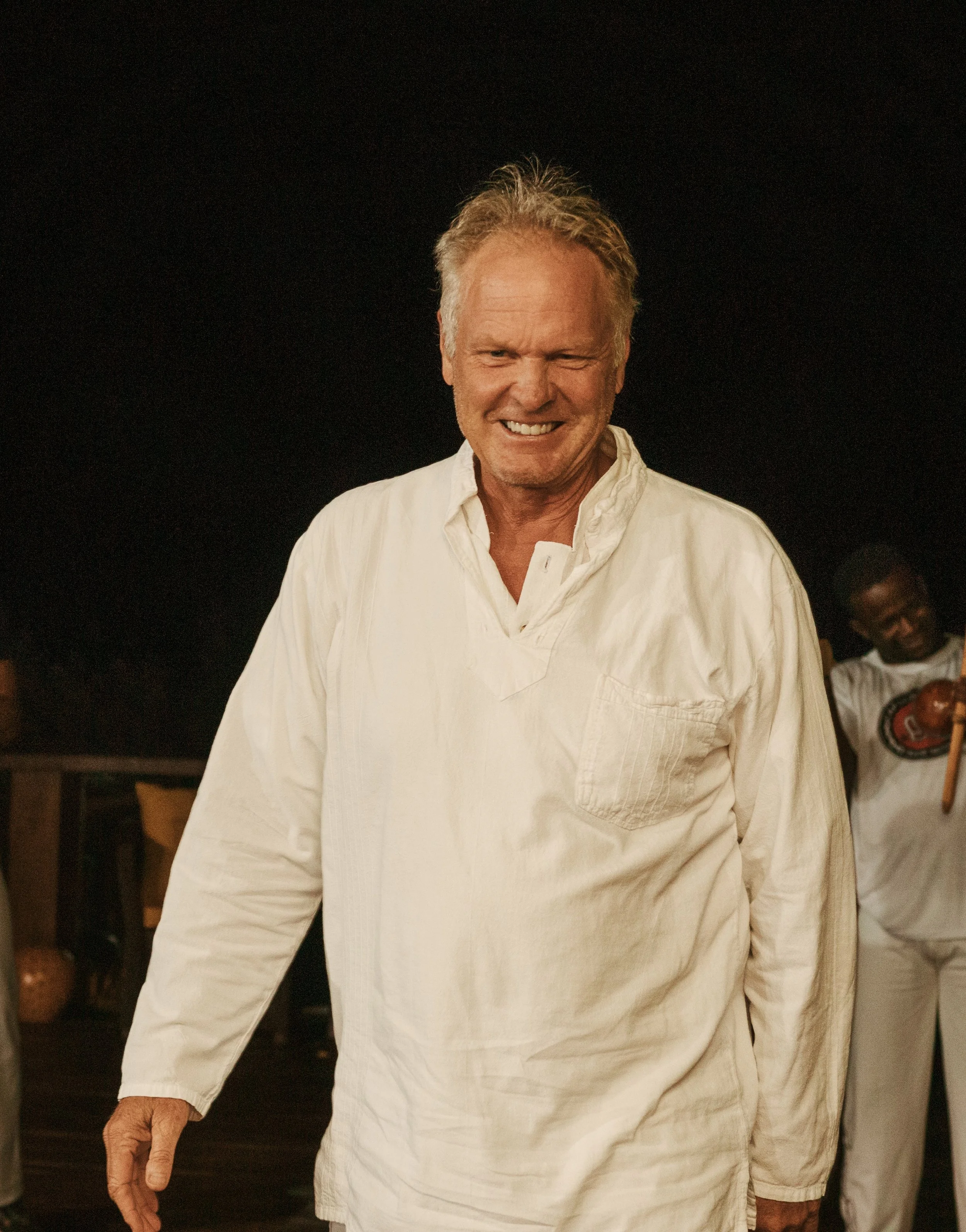 A smiling man with gray hair wearing a white long-sleeve shirt, standing indoors at night.