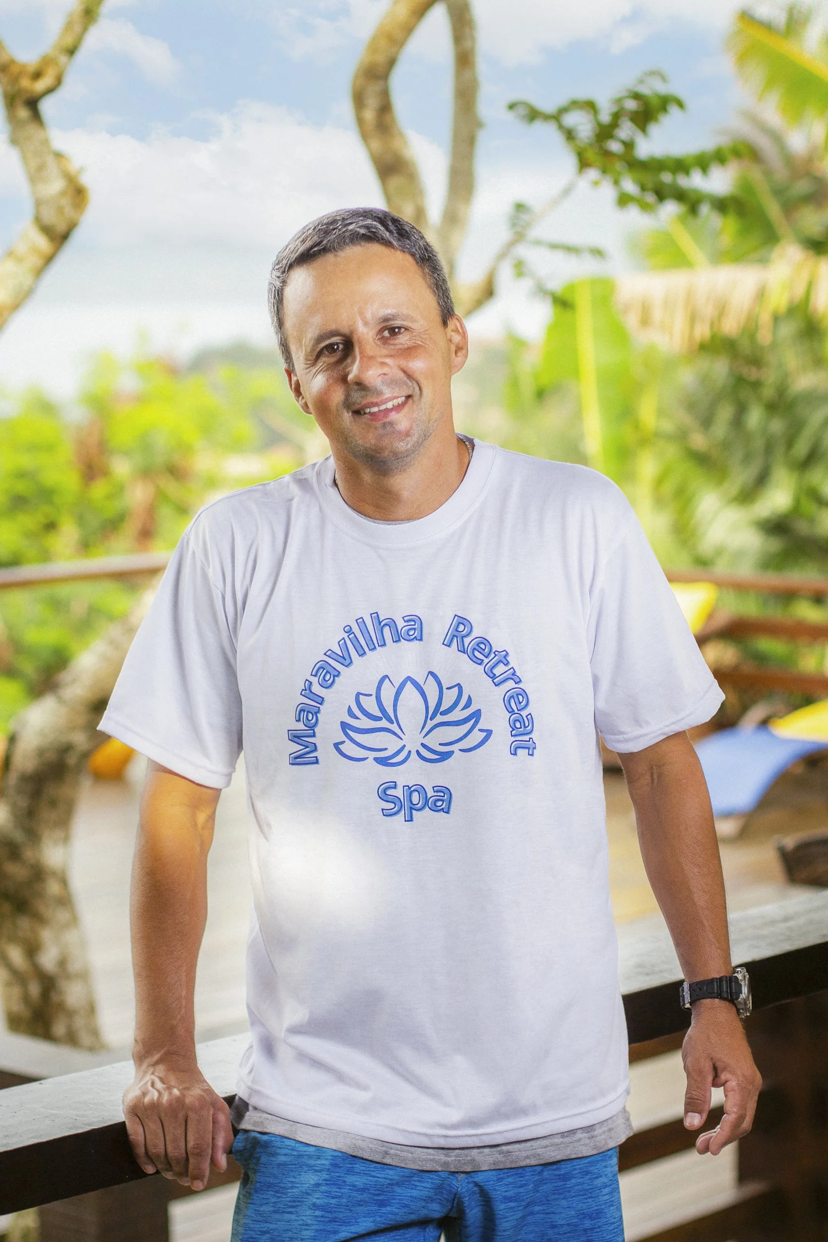 A man smiling and leaning on a wooden railing outdoors, wearing a white T-shirt with a blue floral logo and the text "Maravilha Retreat Spa." There are trees and greenery in the background.