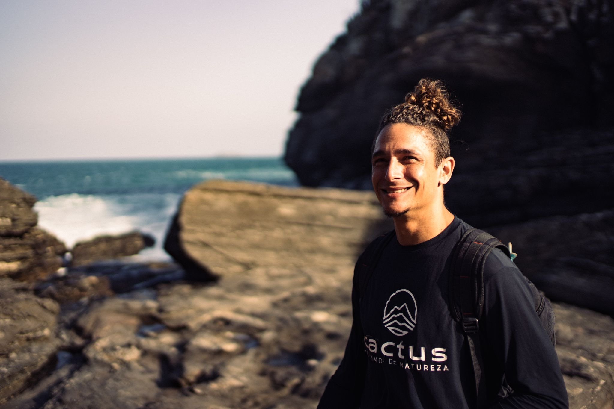 Smiling man with curly hair tied in a bun, wearing a black shirt and backpack, standing on a rocky seaside with cliffs and the ocean in the background.