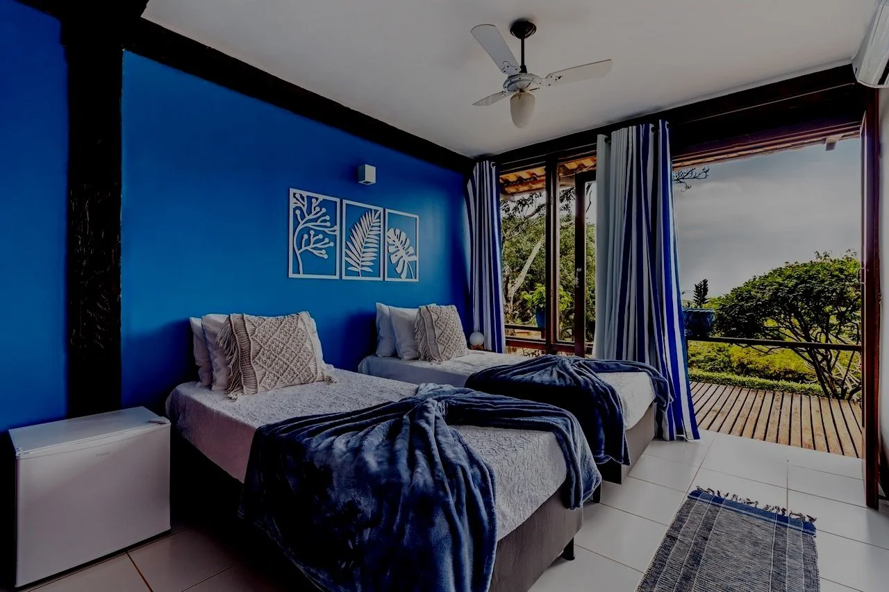 Bedroom with two beds, blue accent wall with framed leaf artwork, open balcony door showing outdoor greenery, white ceiling fan, striped curtains, small white refrigerator, and a gray rug.