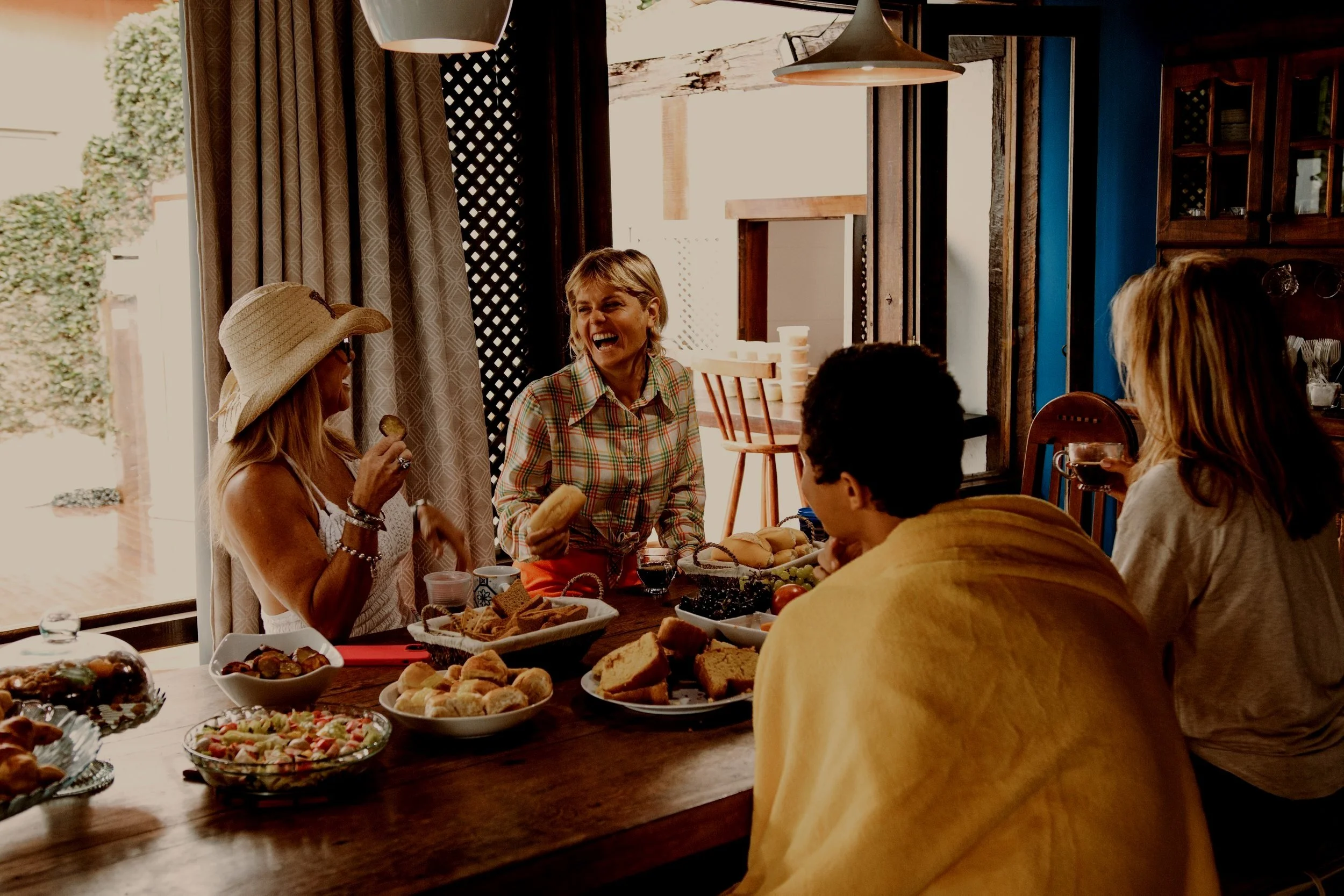 A group of five people enjoying a meal together at a dining table in a cozy, sunlit dining room. They are smiling and laughing, with plates of food including bread, salads, and desserts on the table.