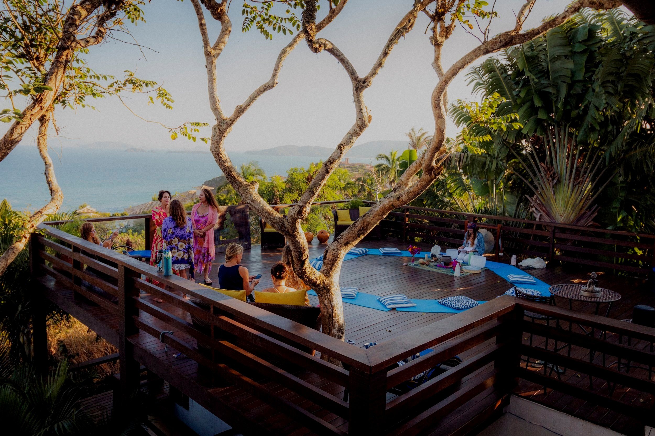 A group of women gathering on a wooden deck with ocean view, some sitting on yellow cushions, others standing and talking, surrounded by tropical trees and plants, with a yoga or meditation setup in the background.