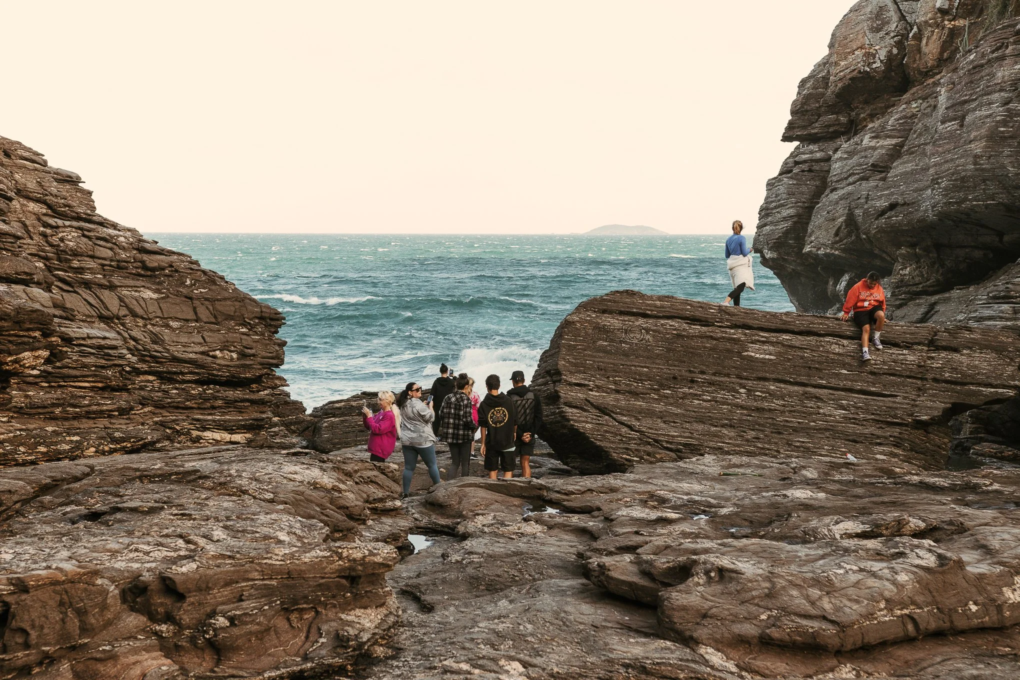 Group of people exploring rocky coastal area with waves and ocean in background.