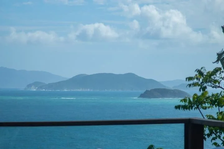 View of the ocean with distant mountainous islands under a partly cloudy sky, with a balcony railing in the foreground and greenery on the right.