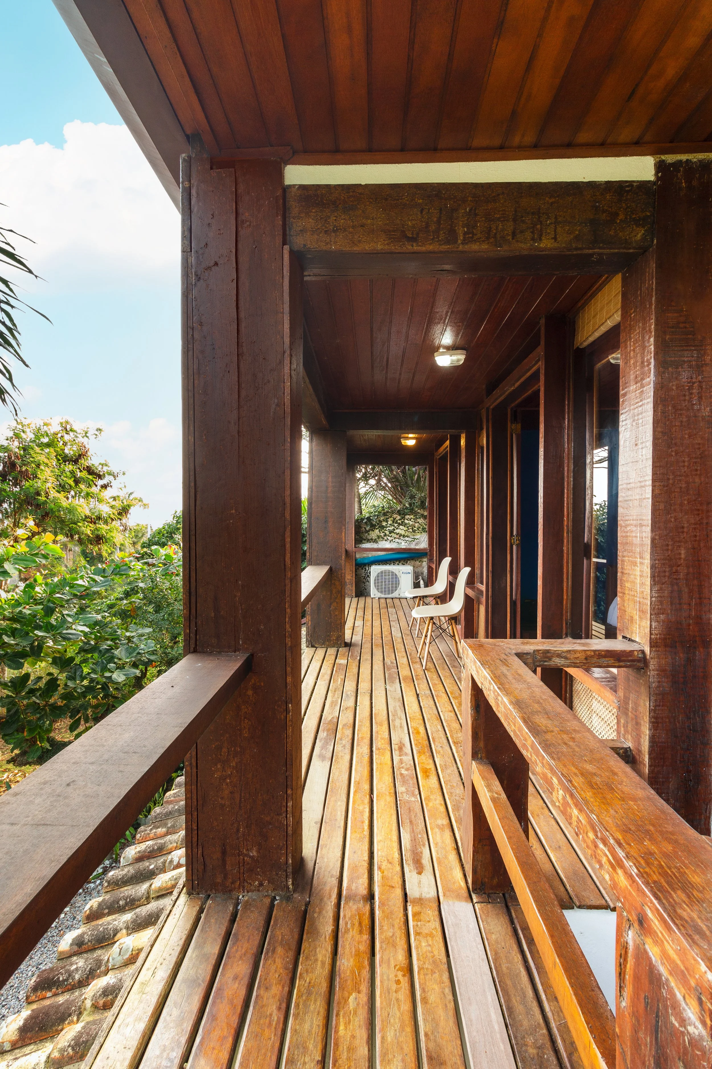 Wooden balcony with two white chairs, overlooking greenery with blue sky and clouds in the background.