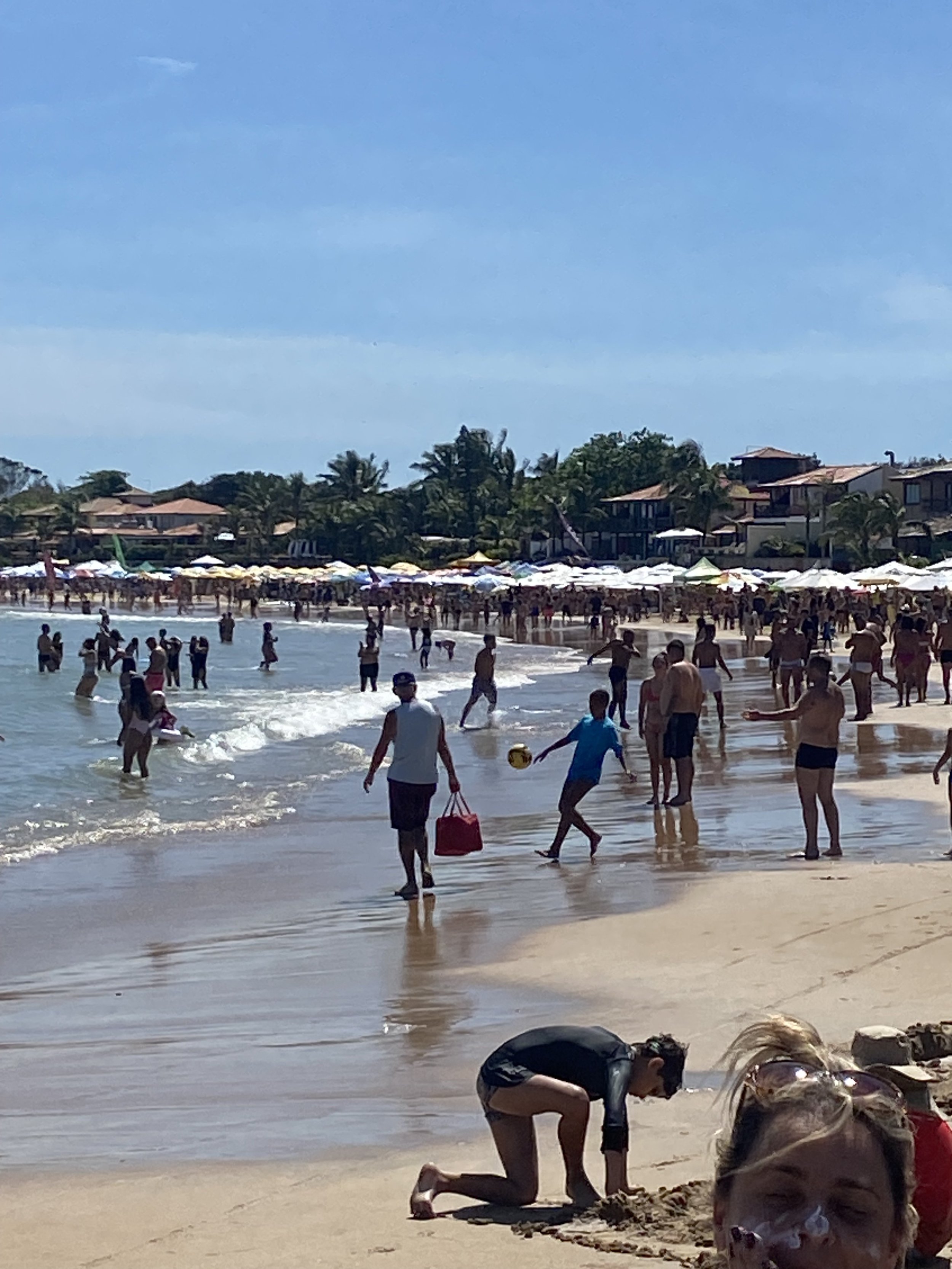 Crowded beach scene with people swimming, walking, and playing in the sand under a clear blue sky. Beach umbrellas and houses are visible in the background.