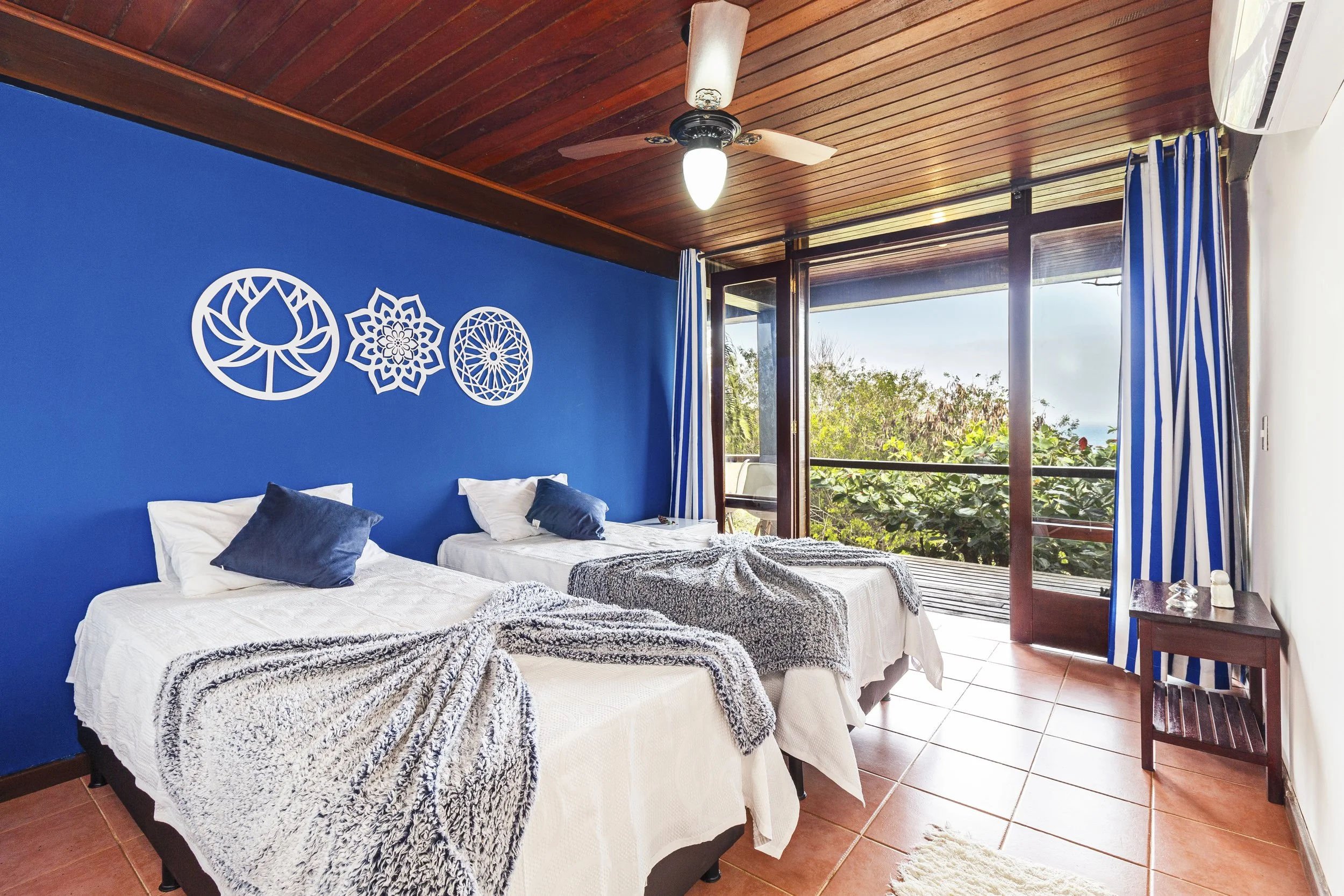A bedroom with two single beds, white bedding, and navy blue pillows. The wall behind the beds is painted blue and has three decorative wall hangings. There is a wooden ceiling fan, a sliding glass door leading to a balcony, and blue and white stripe