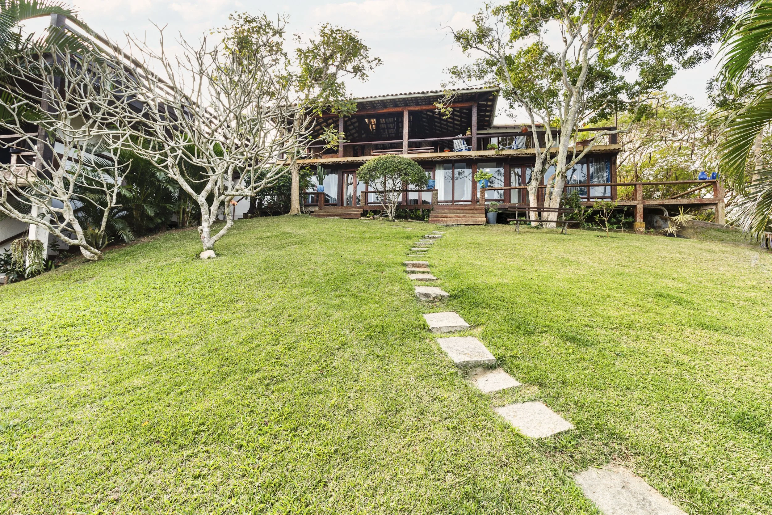 A two-story house with large glass windows and a wooden deck, surrounded by trees and a grassy lawn, with a stone pathway leading to the house.