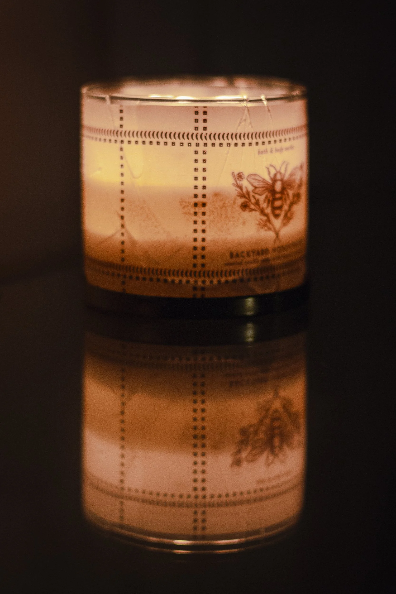 A lit candle in a glass container with botanical and bee illustrations, and measurement markings, casting a warm glow on a black surface with the candle's reflection visible.