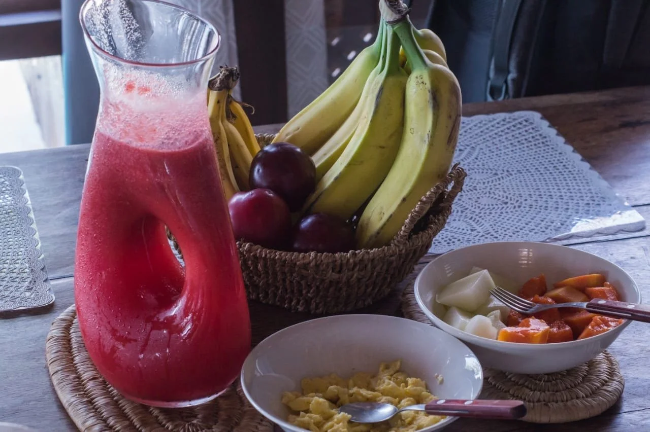 A table set with a basket of bananas and apples, a pitcher of pink fruit juice, and bowls of cut fruit including melons and papaya, with a spoon in each bowl.