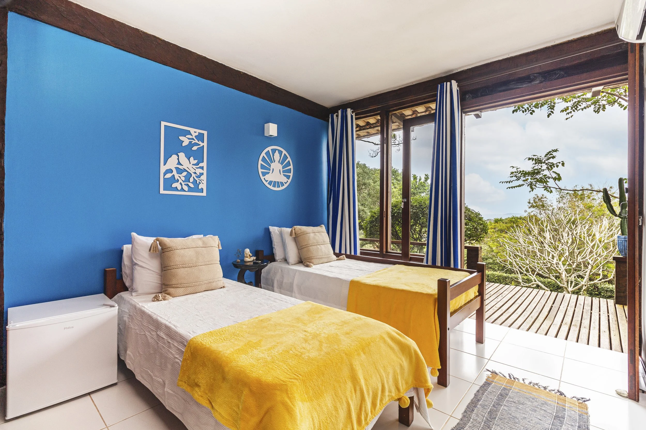 A bright bedroom with two single beds, blue accent wall, yellow blankets, bright view outside, sliding glass doors, and outdoor deck.