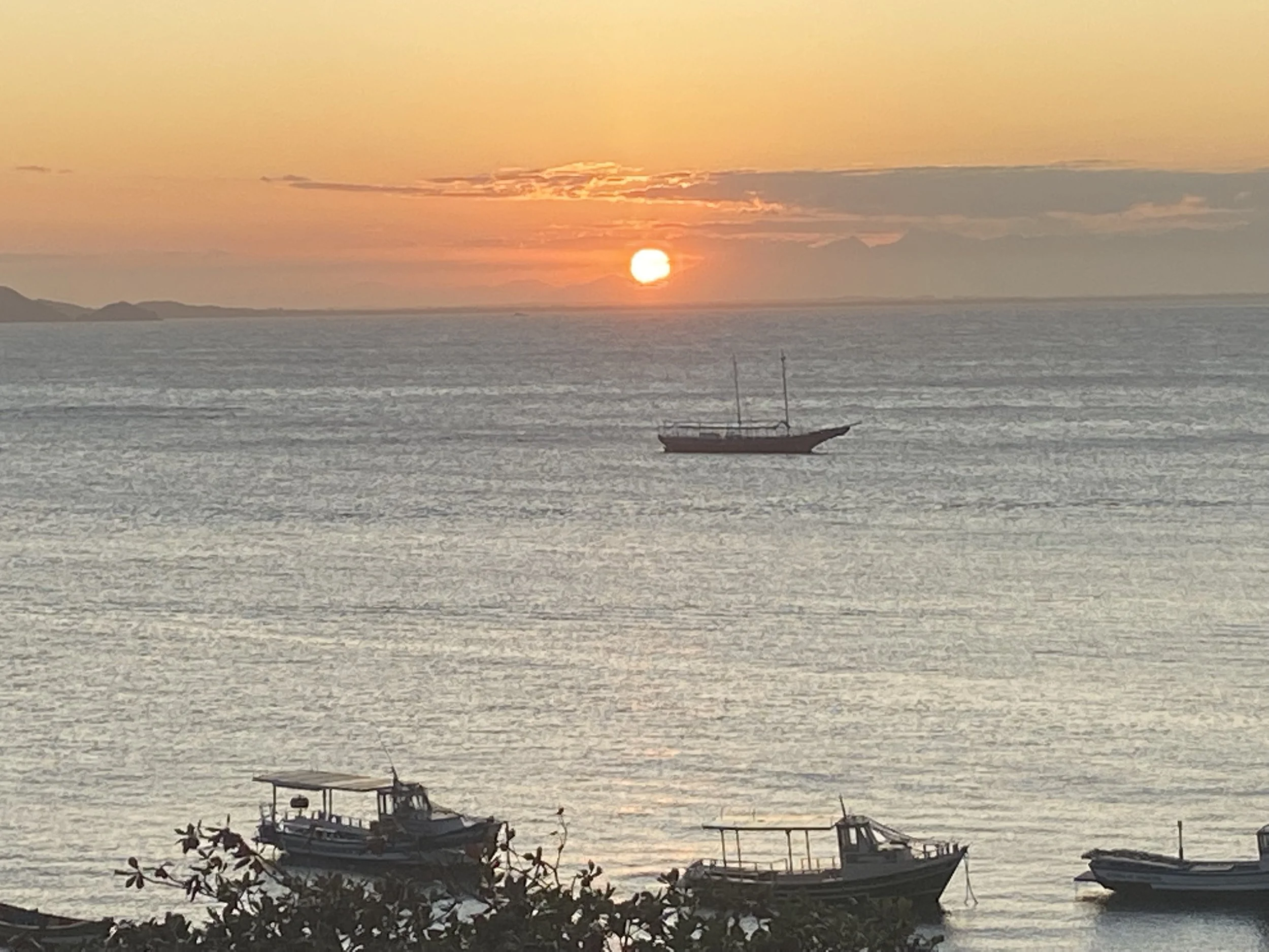 Sunset over the ocean with a sailboat in the distance and several boats docked at the shoreline in the foreground.