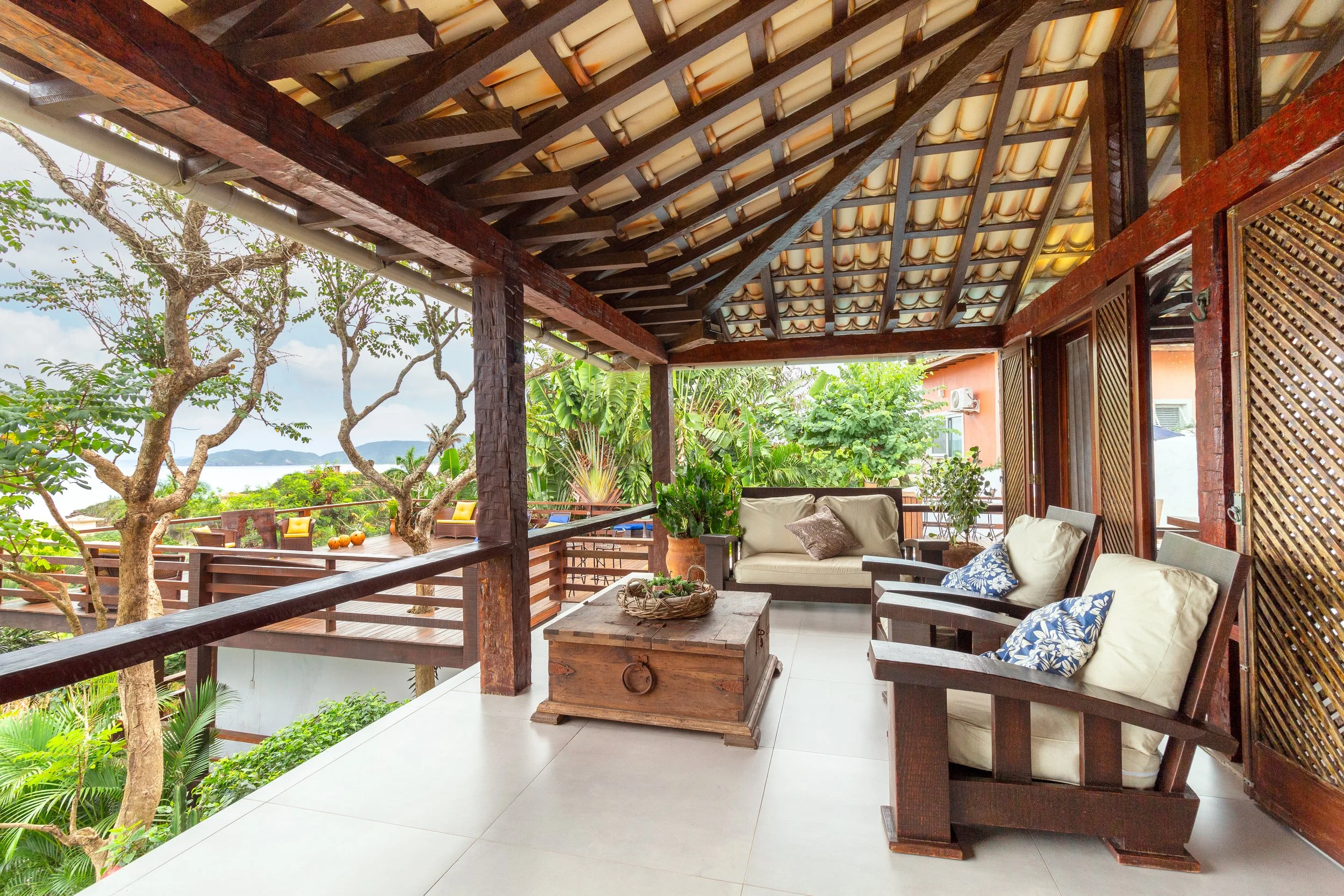 Balcony with wooden furniture, potted plants, and lush greenery with trees, overlooking the ocean during daytime.