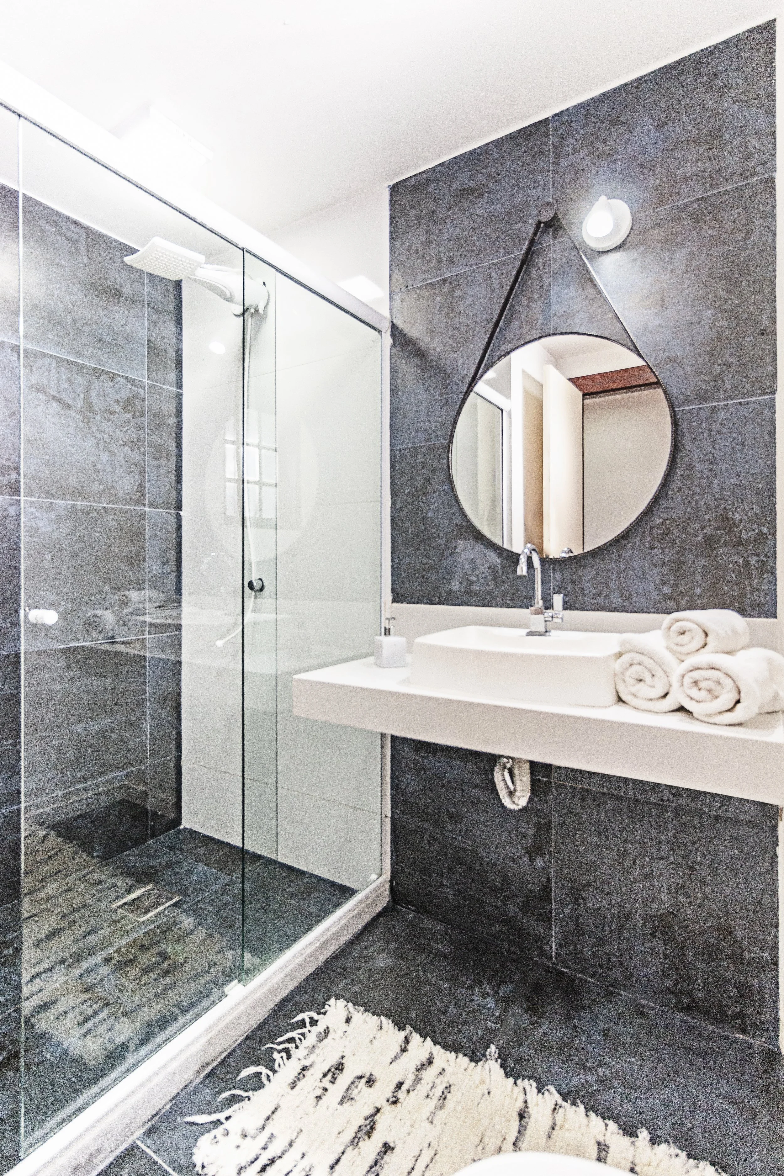 Modern bathroom with dark gray tiled walls and floor, a glass-enclosed shower, a white countertop with a vessel sink, a round mirror, rolled white towels, a small soap dispenser, and a textured white rug.
