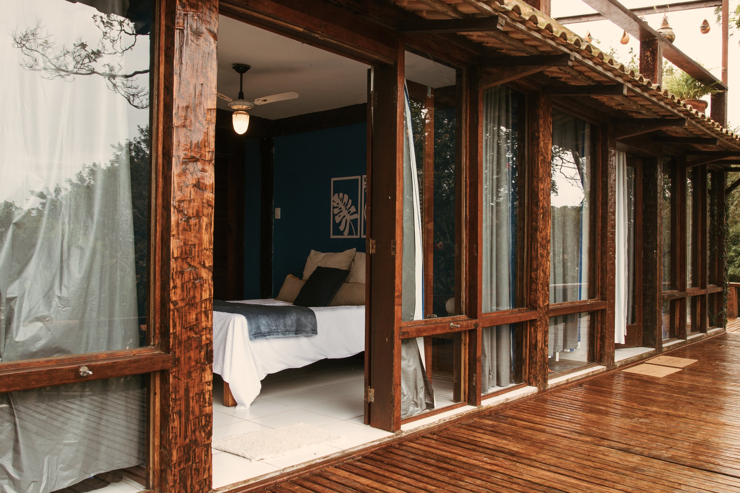 View of a rustic wooden cabin with glass sliding doors, one of which is open, revealing a bedroom with a bed, pillows, and wall art, on a wooden deck during daytime.