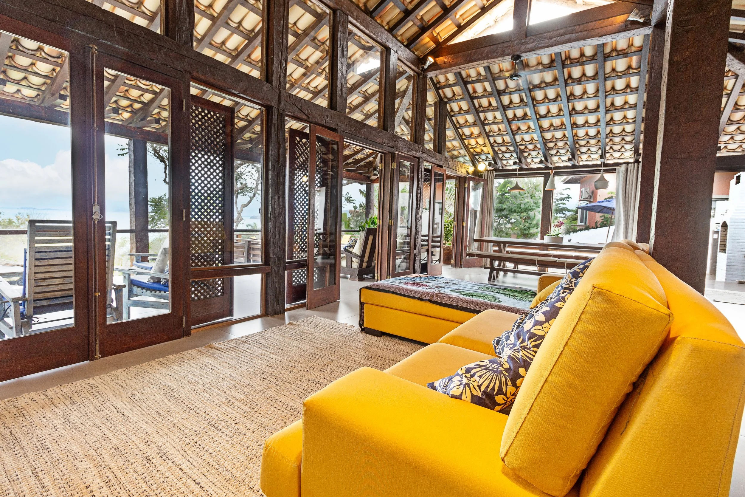 Living room with yellow sectional sofa, blue patterned pillows, wooden beams, glass doors, outdoor patio with chairs and greenery visible outside.