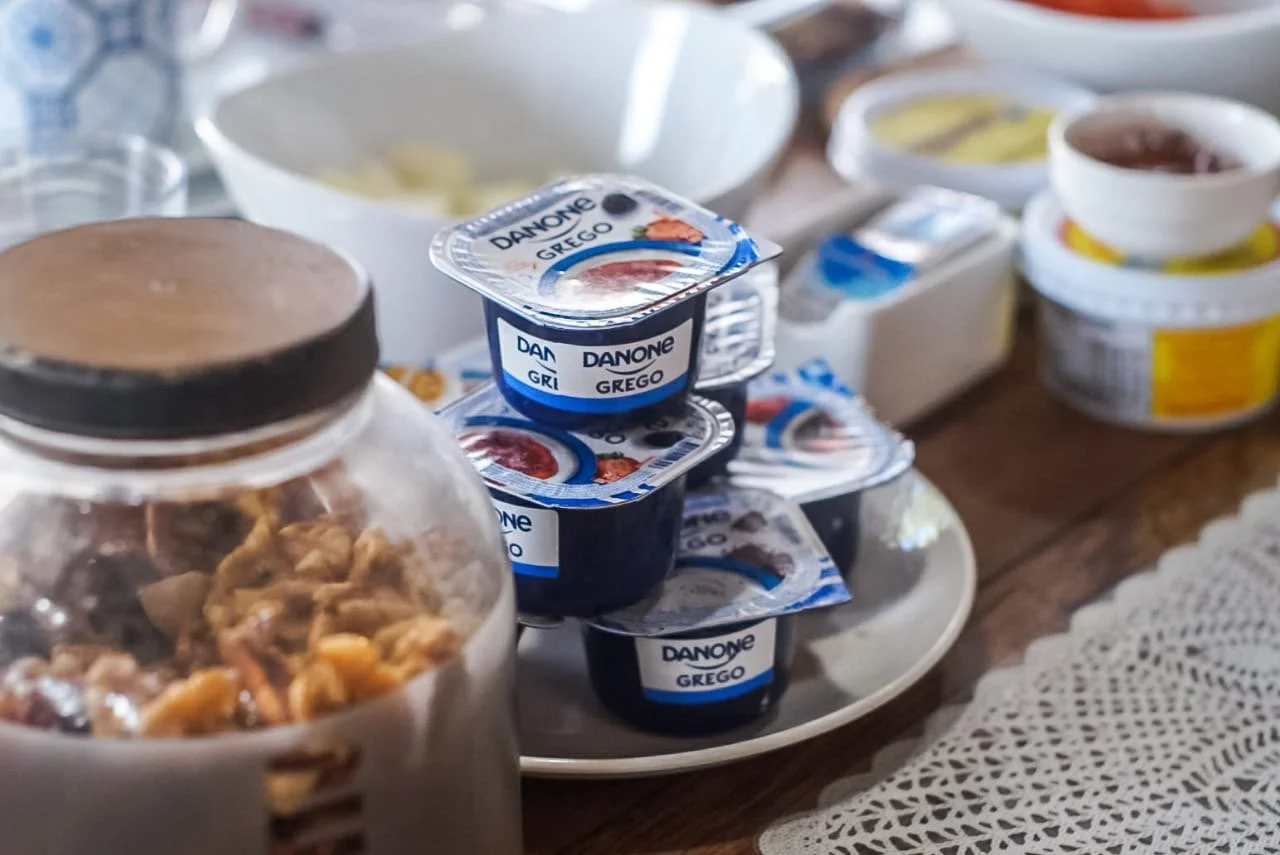 A table with multiple containers of Greek yogurt, a jar of cereal, and a bowl of cereal on a lace tablecloth.