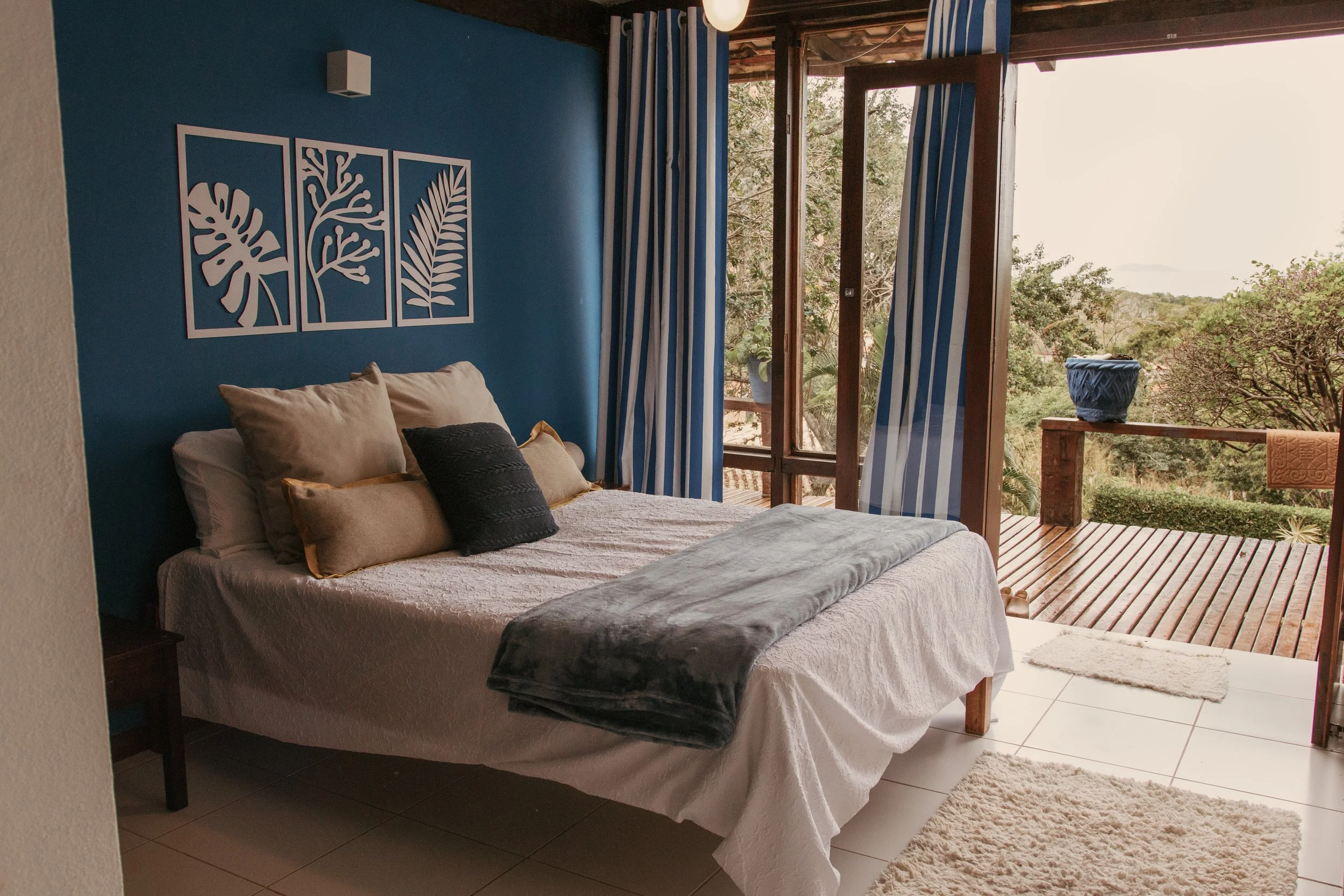 A bedroom with a bed featuring beige, black, and gold pillows, a white textured blanket, and a gray throw. The room has a blue accent wall with white leaf art, a small black nightstand, and a balcony with striped blue and white curtains, a wooden dec