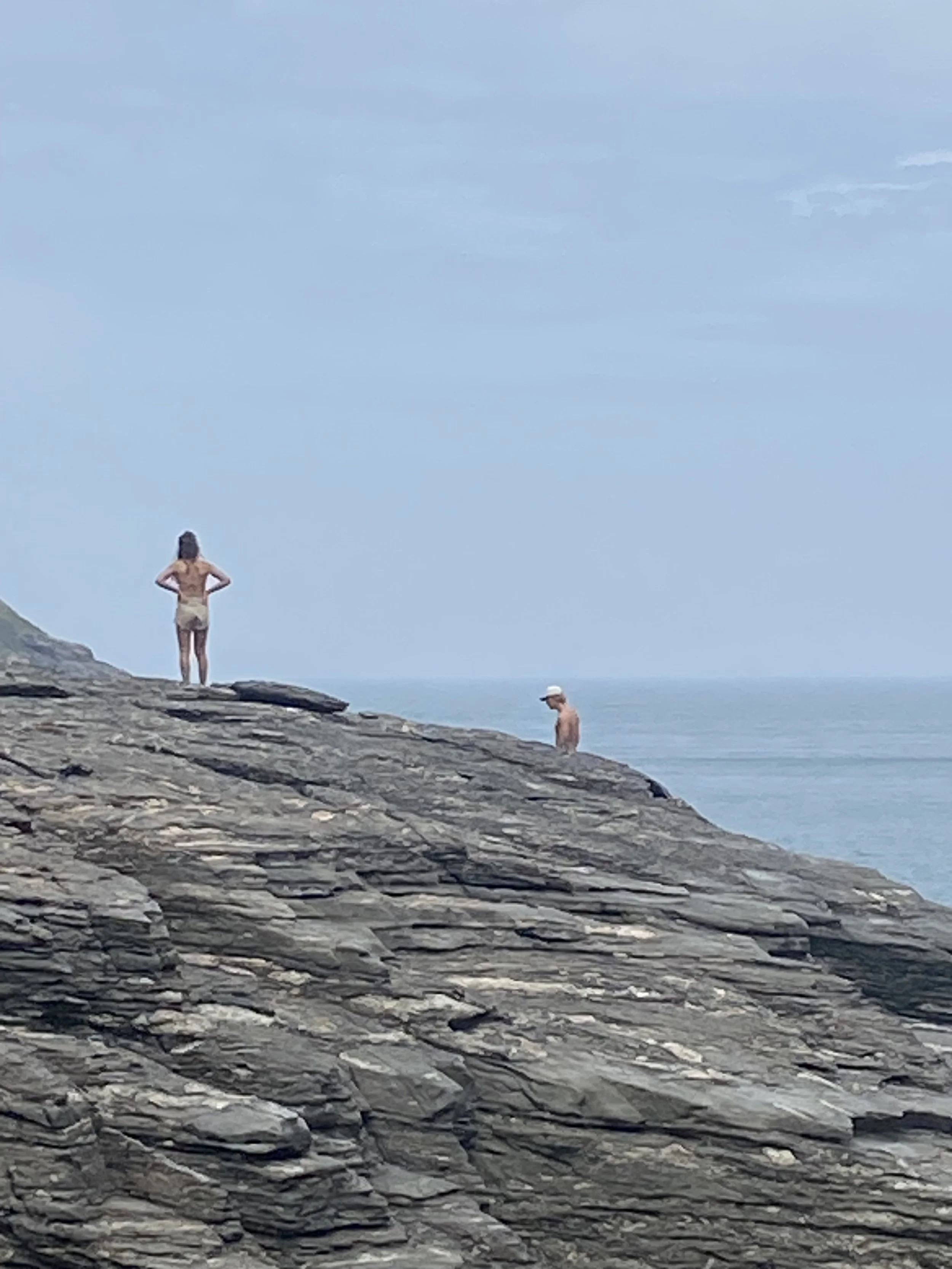 Two people on a rocky shoreline, one woman standing with hands on hips and one man sitting, overlooking the ocean under a cloudy sky.