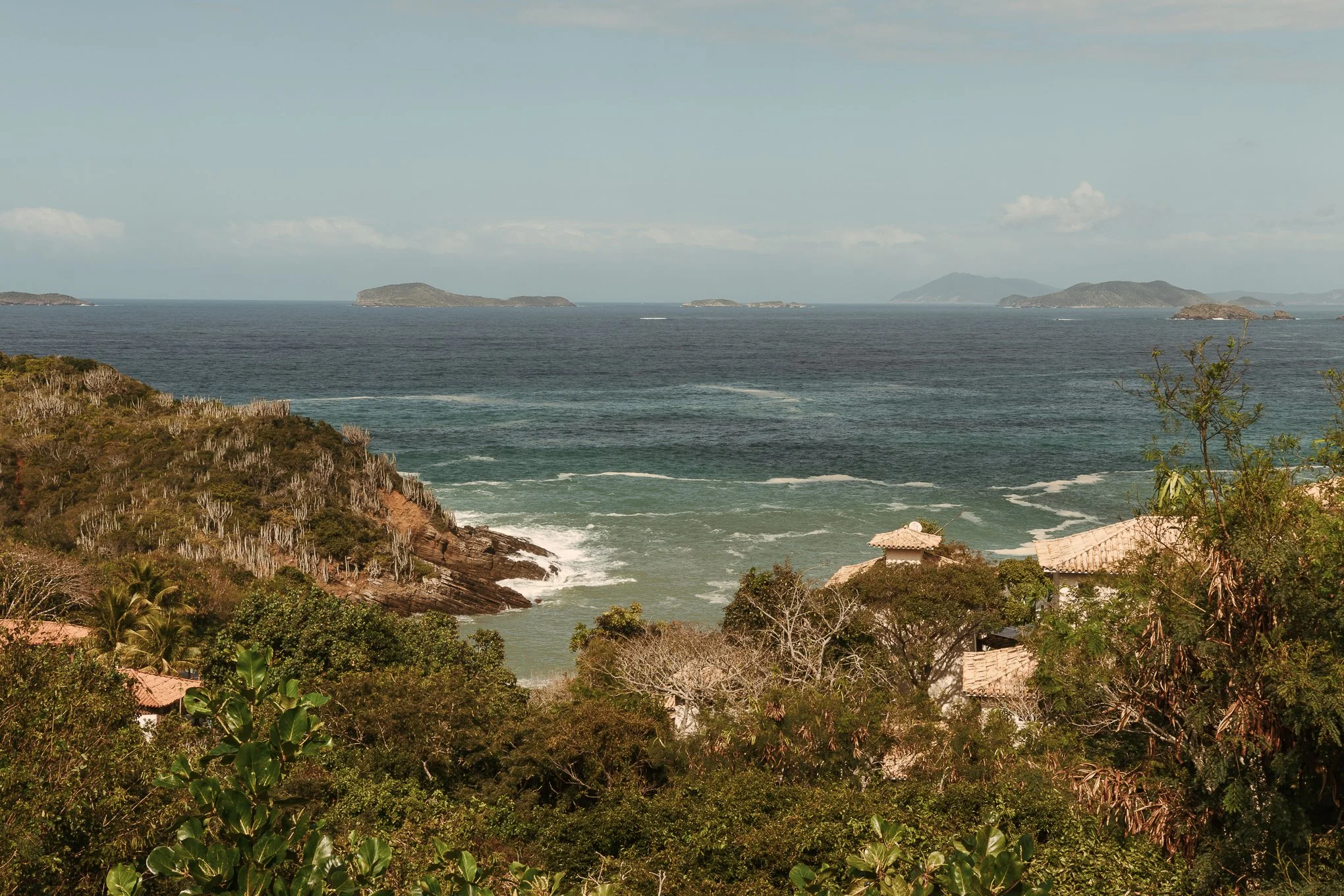 Coastal landscape with trees and houses in the foreground, rocky cliffs, and a body of water with islands in the background under a blue sky with clouds.