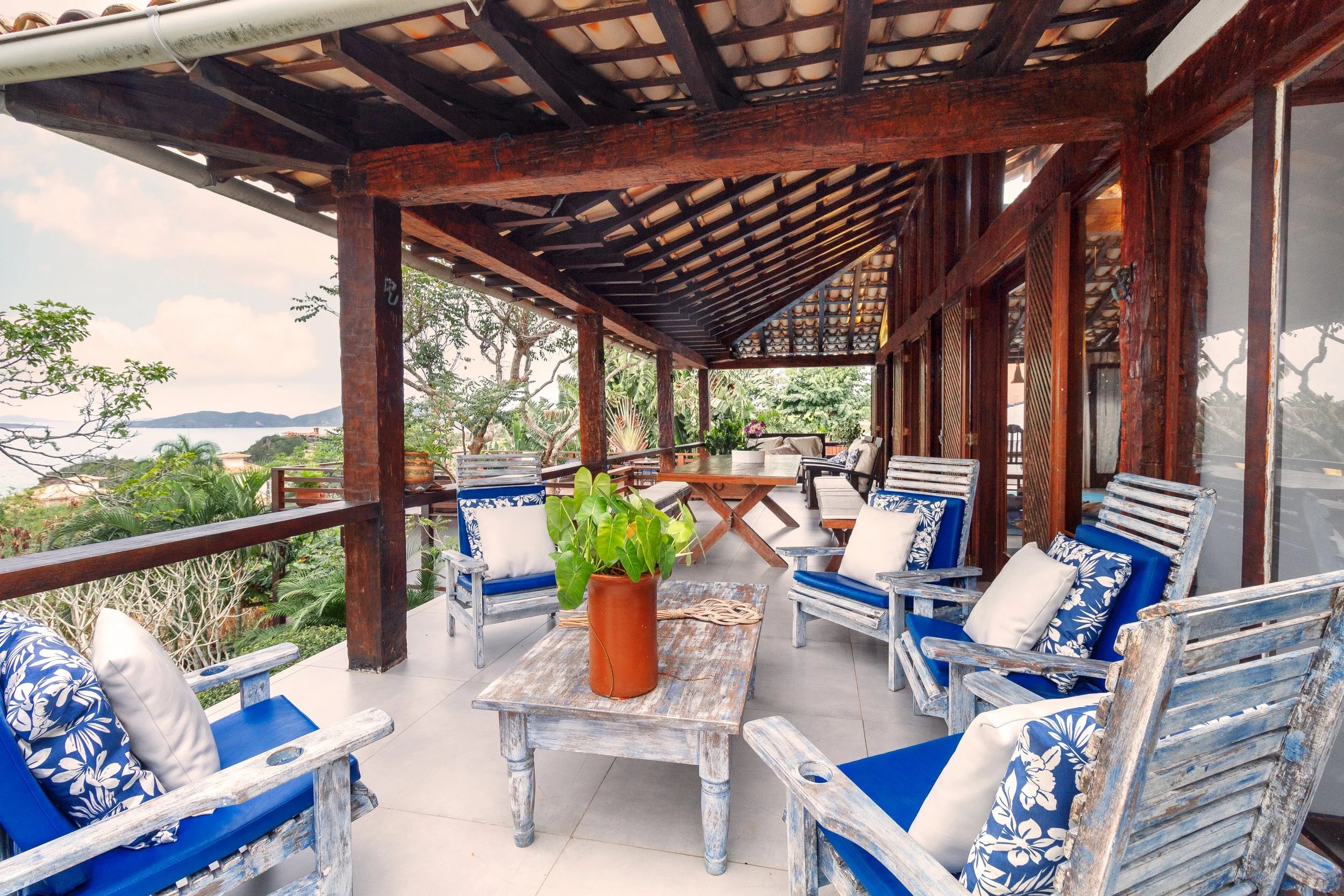 A spacious outdoor balcony with vintage wooden chairs and a table, decorated with cushions and a potted plant, overlooking a lush landscape with trees and a distant body of water.