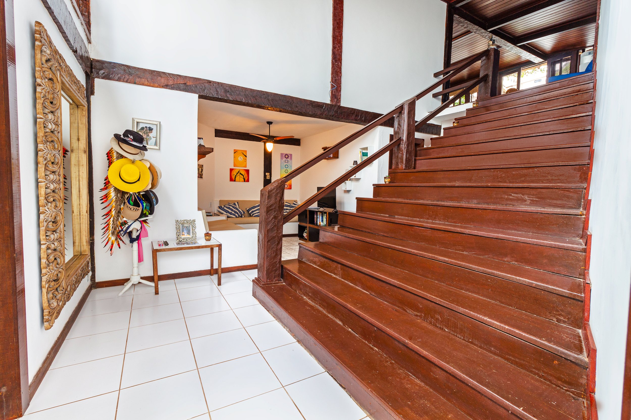 Living room with white tiled floor, wooden staircase, and decor including hats on a stand, a small table, painting on walls, and a built-in couch area.