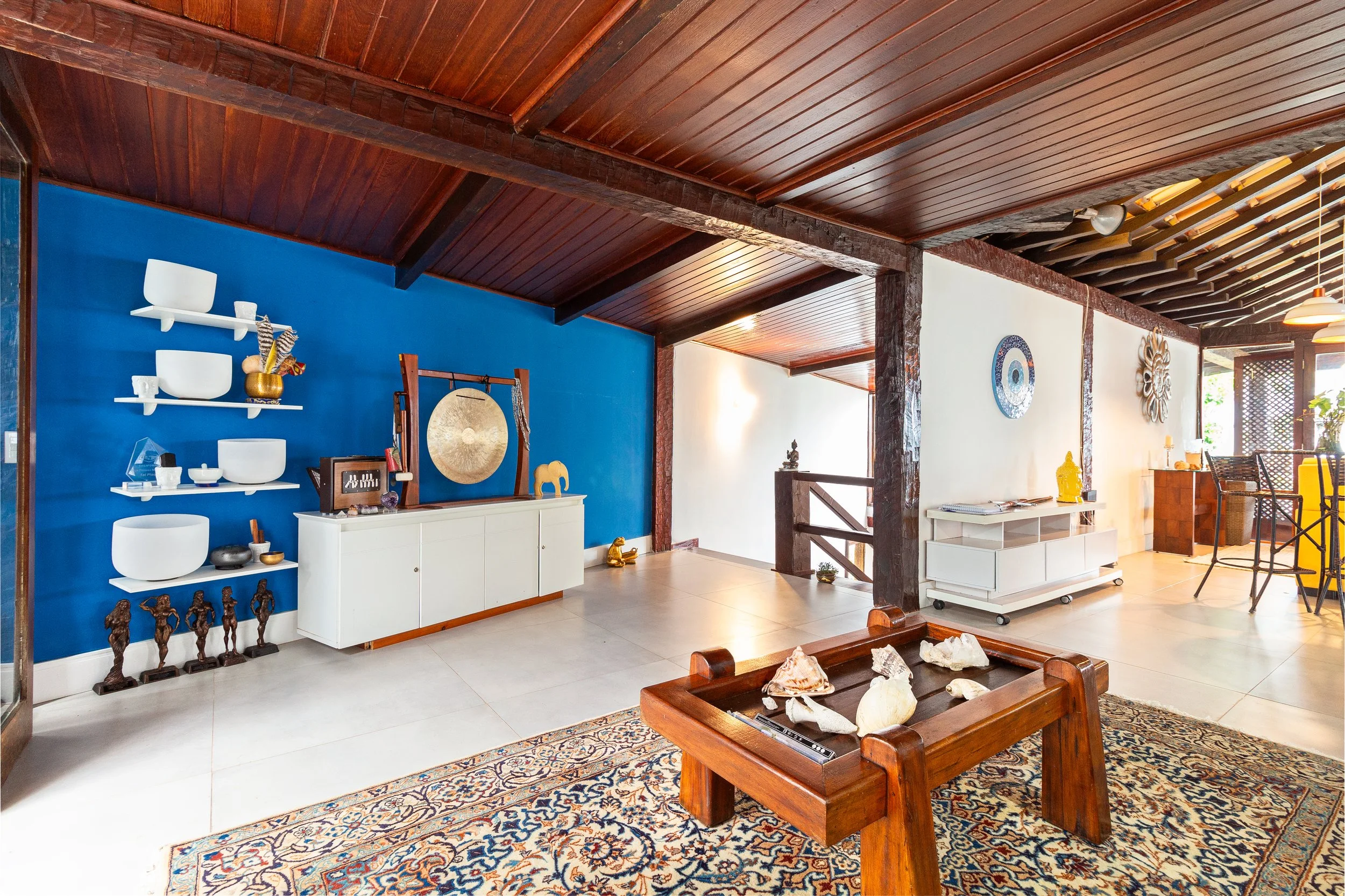 Interior of a living room with a wooden ceiling, white walls, and decorative items including sculptures, a gong, and shells on a wooden table. A blue accent wall is visible with shelves holding various ornaments. There is a white cabinet and a tradit