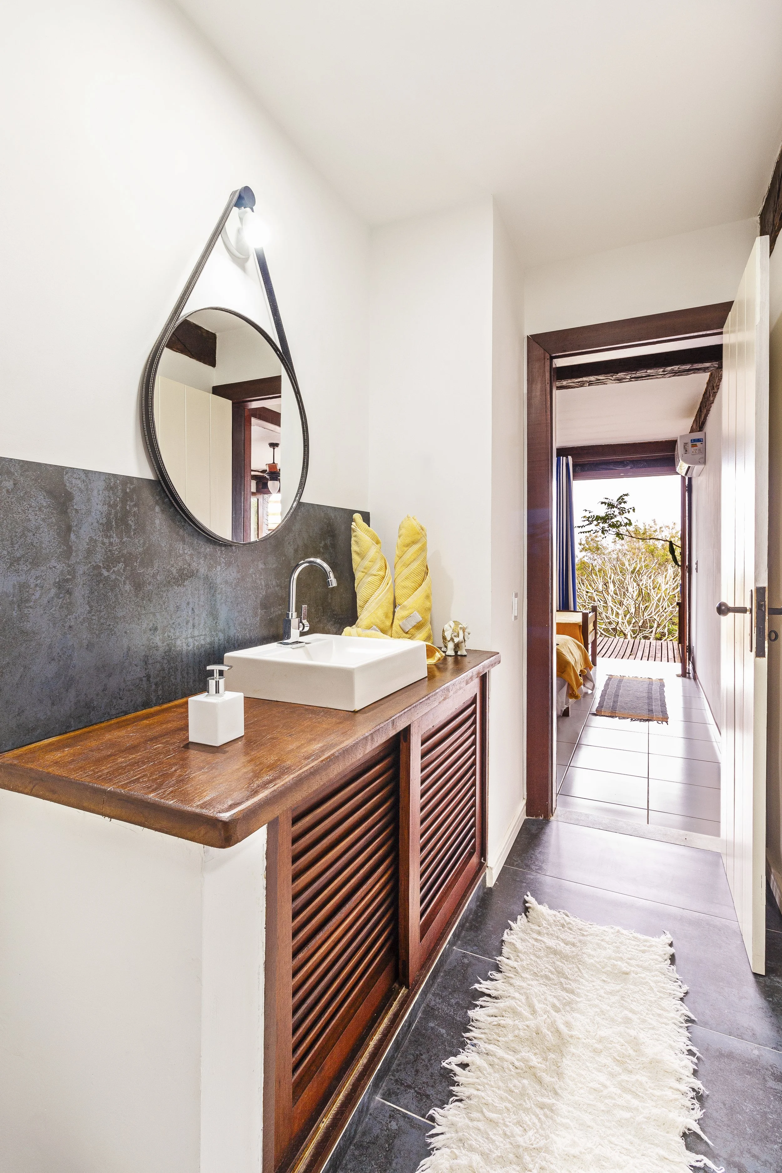 Bathroom with a white sink, wooden cabinet, round mirror, yellow towels, soap dispenser, and a doorway leading to a room with outdoor views.