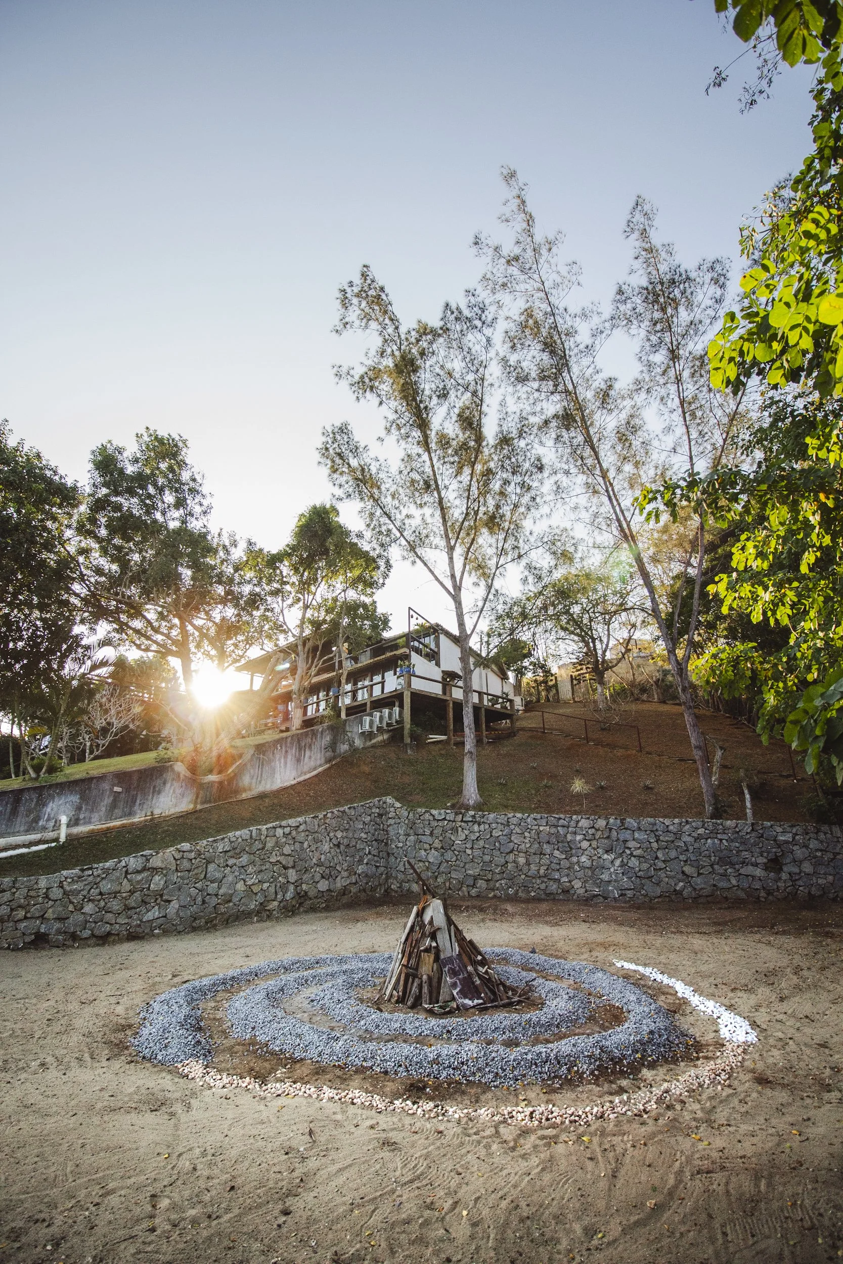 A small outdoor fire pit made of wood logs, surrounded by circular patterns of stones and gravel, with trees and a house in the background during sunset.