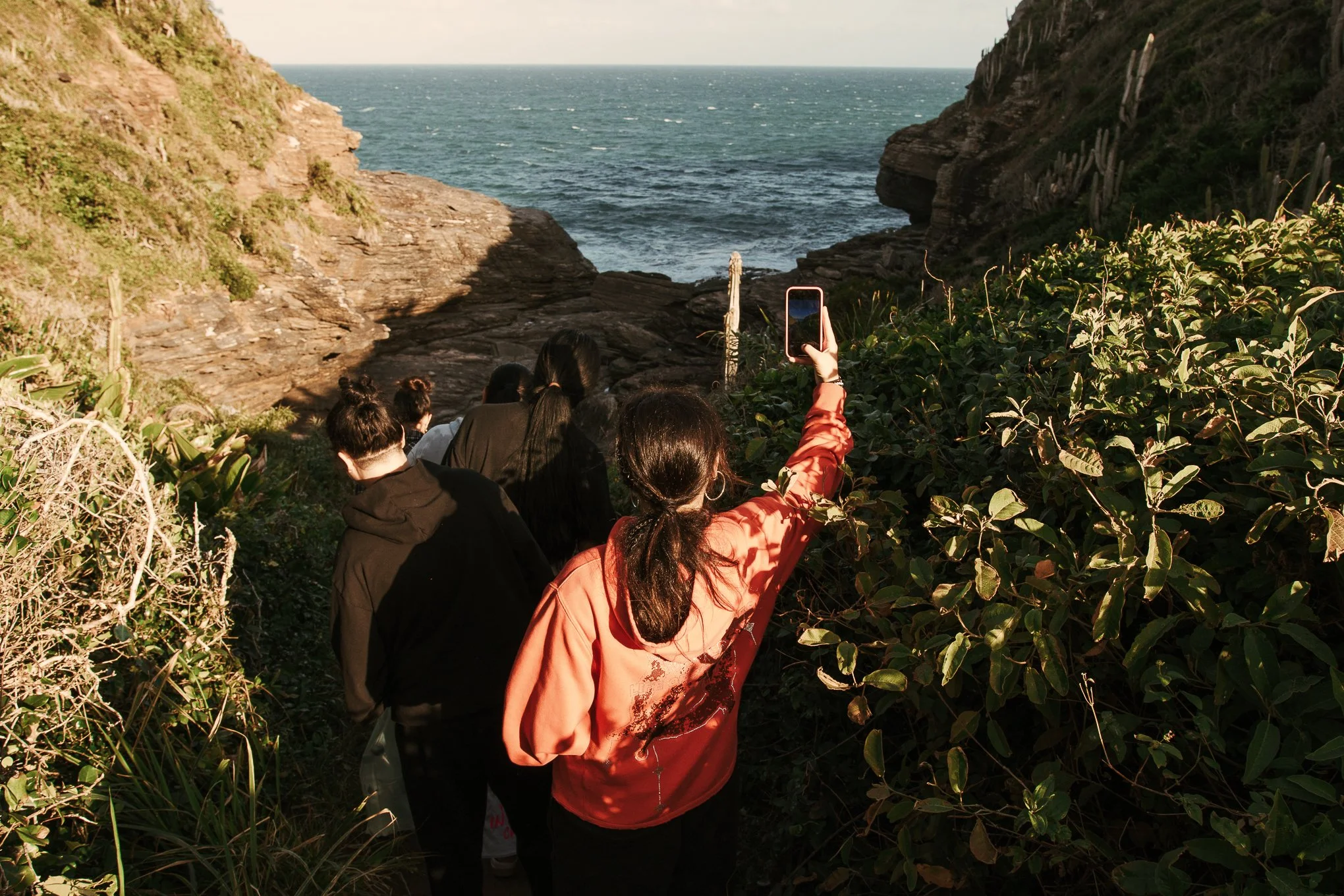 Group of people walking down a rocky trail toward the ocean, with a woman taking a photo with her phone, surrounded by greenery and cliffs.