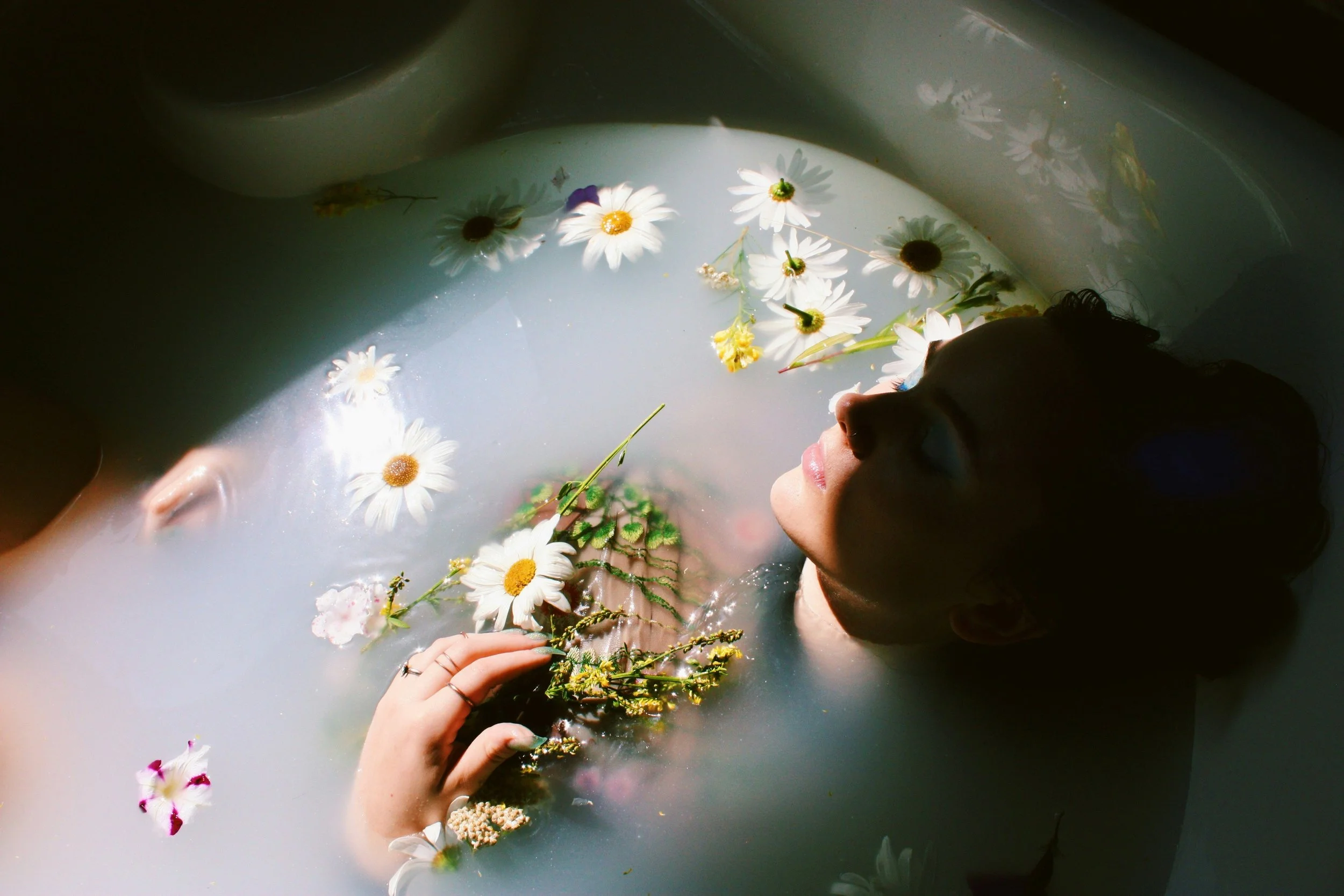 A person with dark hair is lying in a bathtub filled with milky water and floating daisies, holding a small bouquet of flowers.