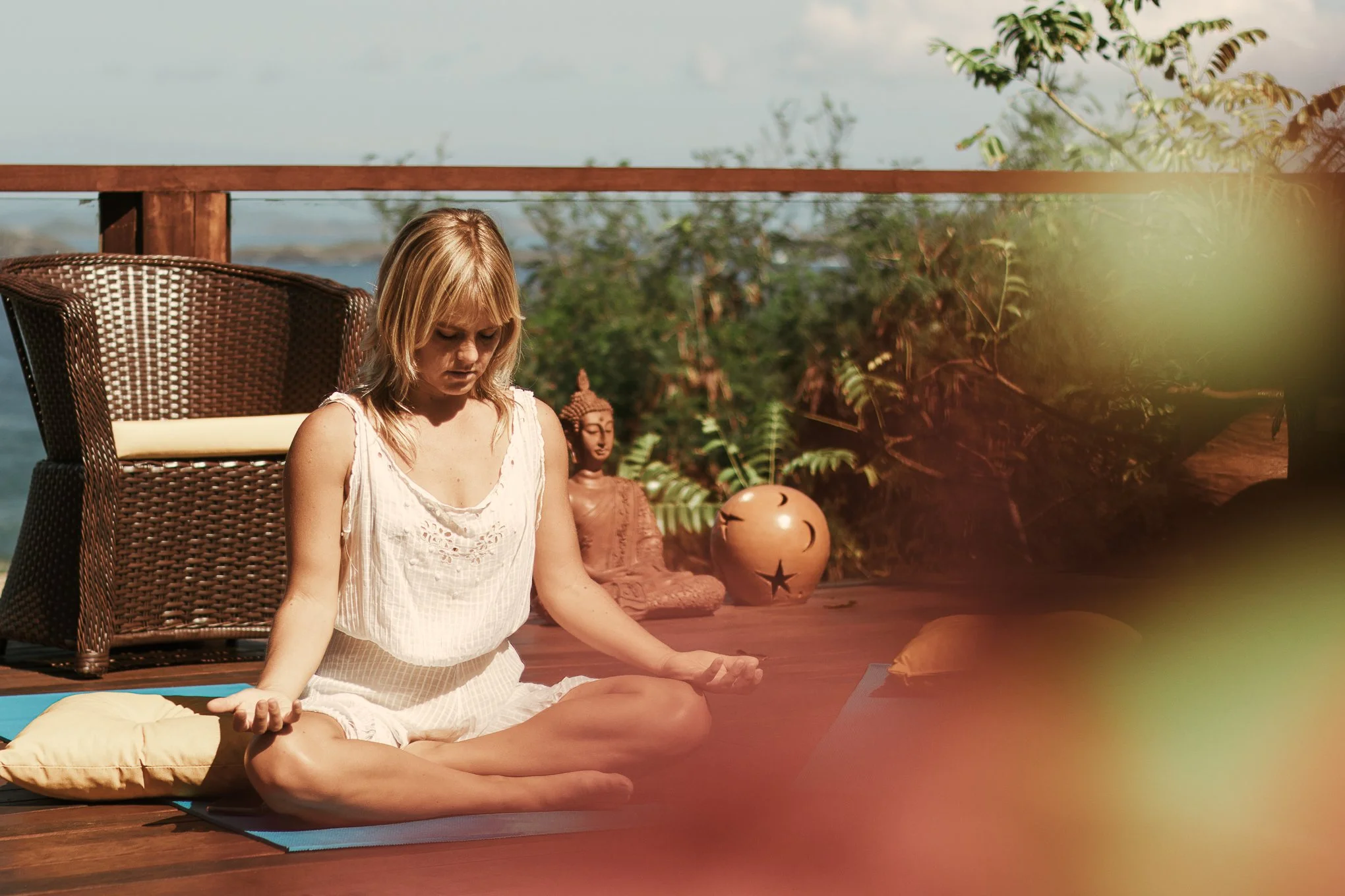 A woman practicing yoga outdoors on a wooden deck, sitting cross-legged with hands resting on knees, surrounded by greenery with a Buddha statue and decorative pot in the background.