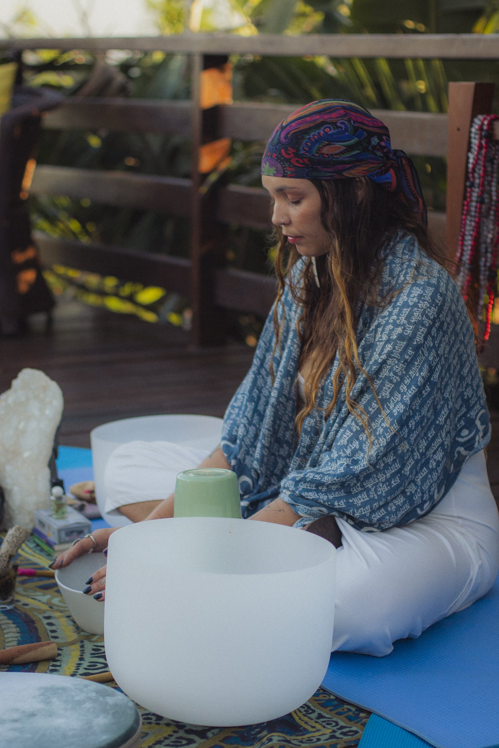 A woman sitting cross-legged on a blue yoga mat outdoors during daytime, with her eyes closed and head slightly bowed, surrounded by spiritual objects and decorative items.