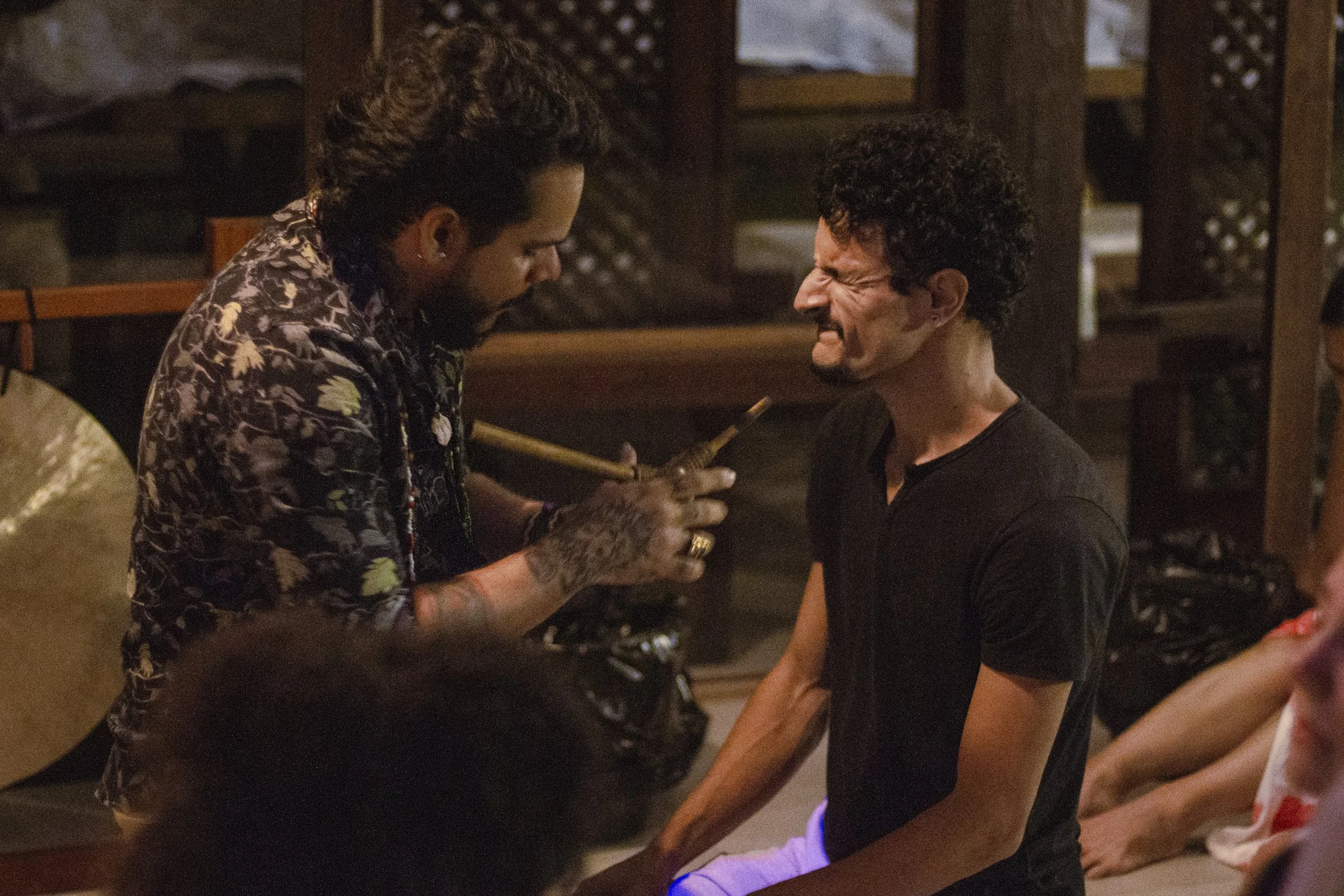 Two men are in a tense moment, one with tattoos and tropical print shirt holding an object close to the face of another man in a black shirt who appears distressed.