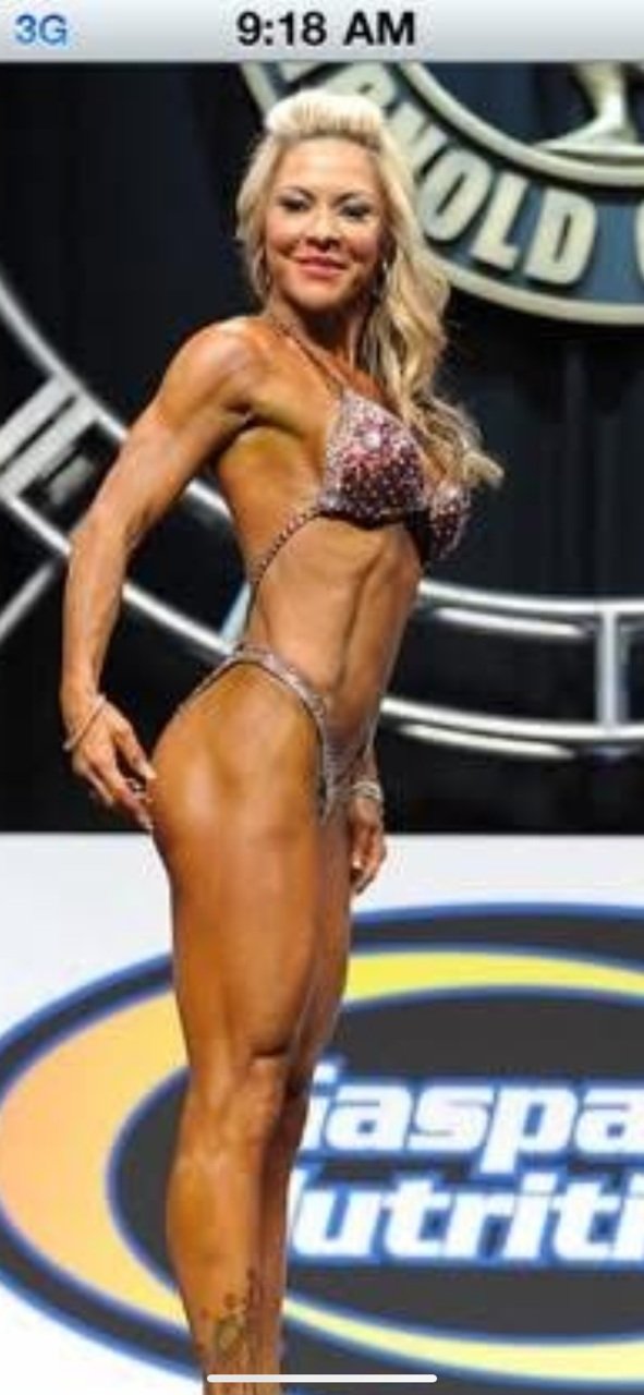 Female bodybuilder posing on stage at a fitness competition, wearing a bikini with a medal around her neck, with a backdrop displaying a logo.