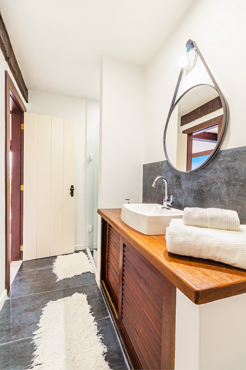 Bathroom with a wooden vanity, white vessel sink, oval mirror, dark gray backsplash, rolled towels, and beige rugs on dark gray tile floor.
