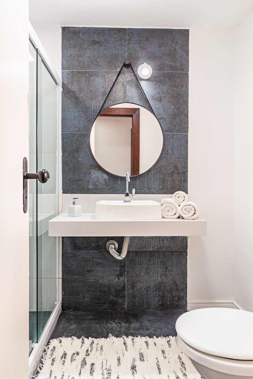 Modern bathroom with black tiled wall, white sink, round mirror, roll towels, and a glass shower door.