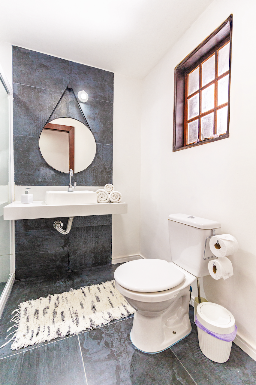Bathroom with white toilet, wall-mounted sink with rolls of white towels, dark tiled wall with round mirror, small mirror, and window with wooden frame.