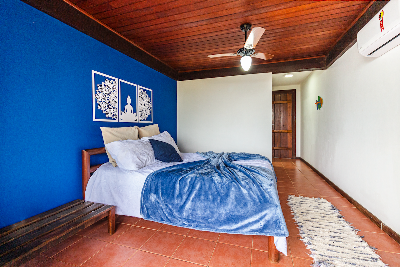 A bedroom with a wooden ceiling and a bed covered in white, beige, and blue blankets against a blue accent wall decorated with white pictures of a Buddhist figure and lotus designs. There is a small wooden bench, a beige and black pillow on the bed, 